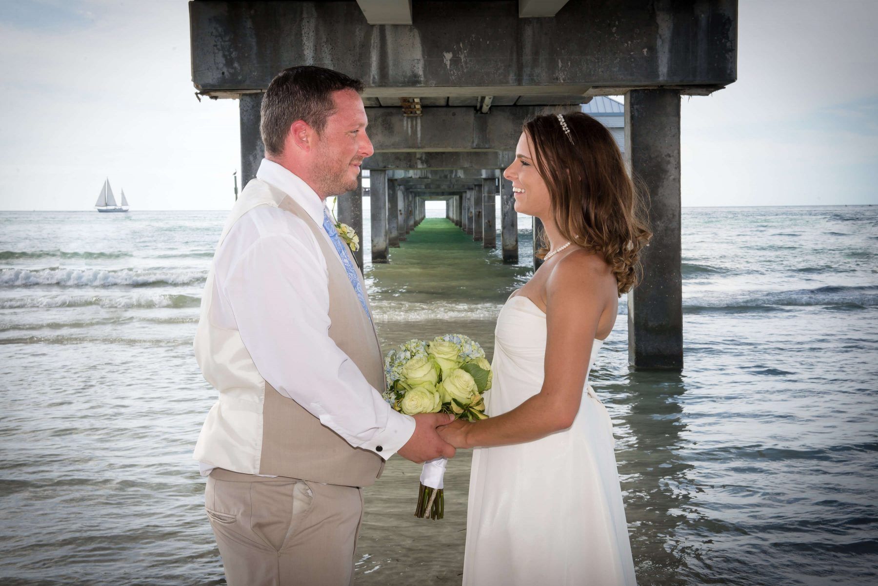 Bride and groom holding hands, smiling under a pier at the beach. Wedding attire and bouquet. Sailboat in the distance.