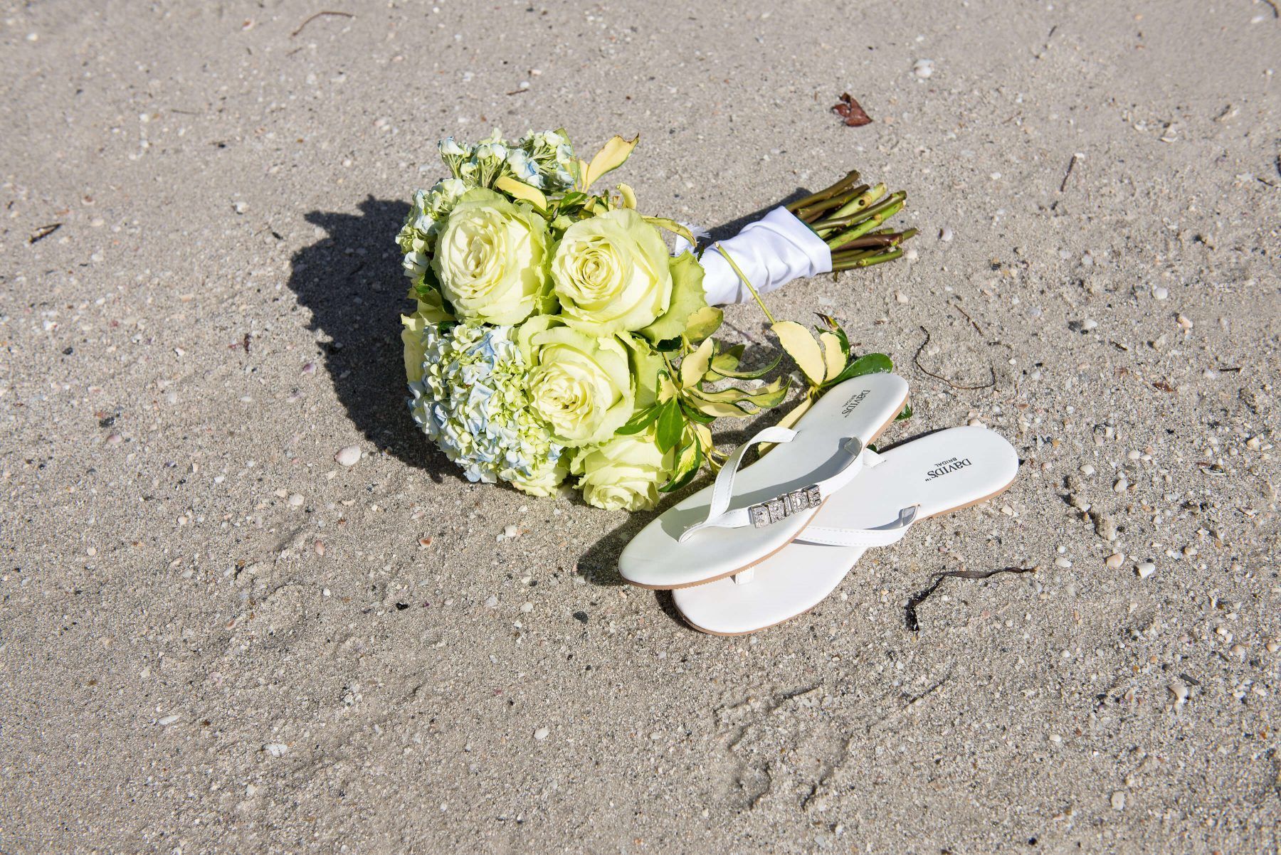 Bouquet of yellow flowers and white sandals on a sandy beach.