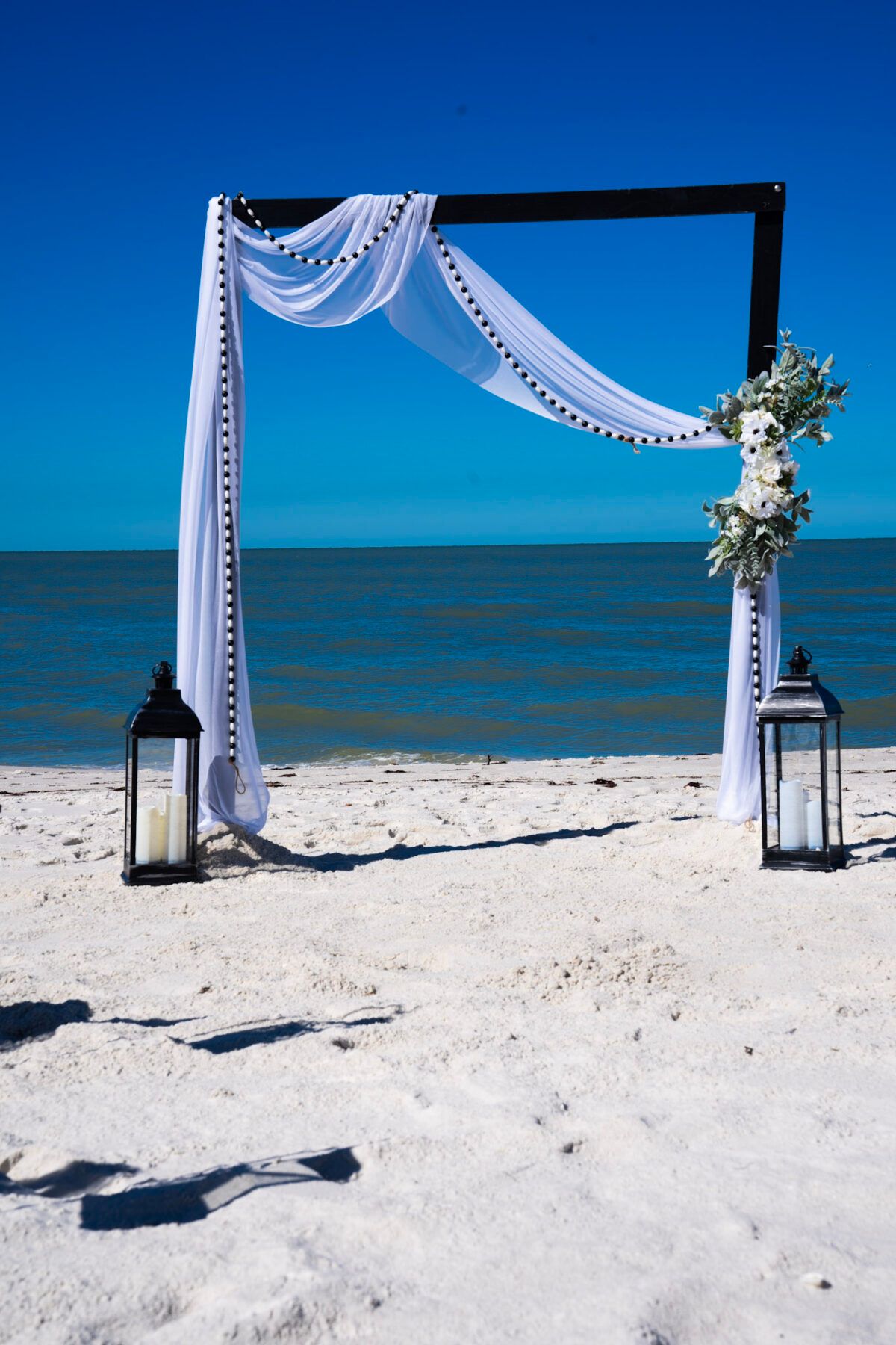 Wedding arch on a white sandy beach with ocean background. Black lanterns flank the arch, draped with white fabric and flowers.
