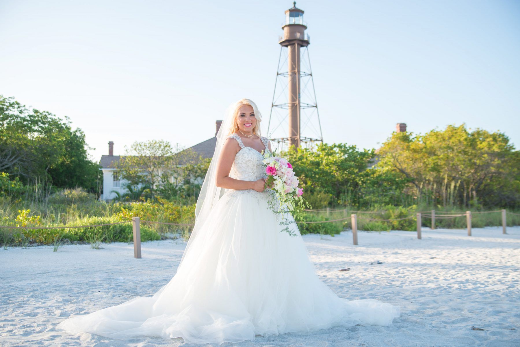 Bride in wedding dress, holding bouquet, poses on beach, with lighthouse in background.
