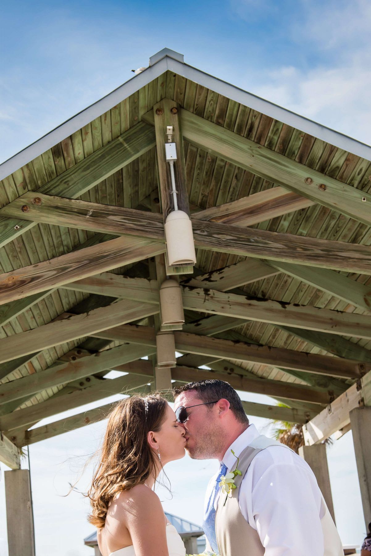 Couple kissing under a wooden gazebo with a blue sky background.