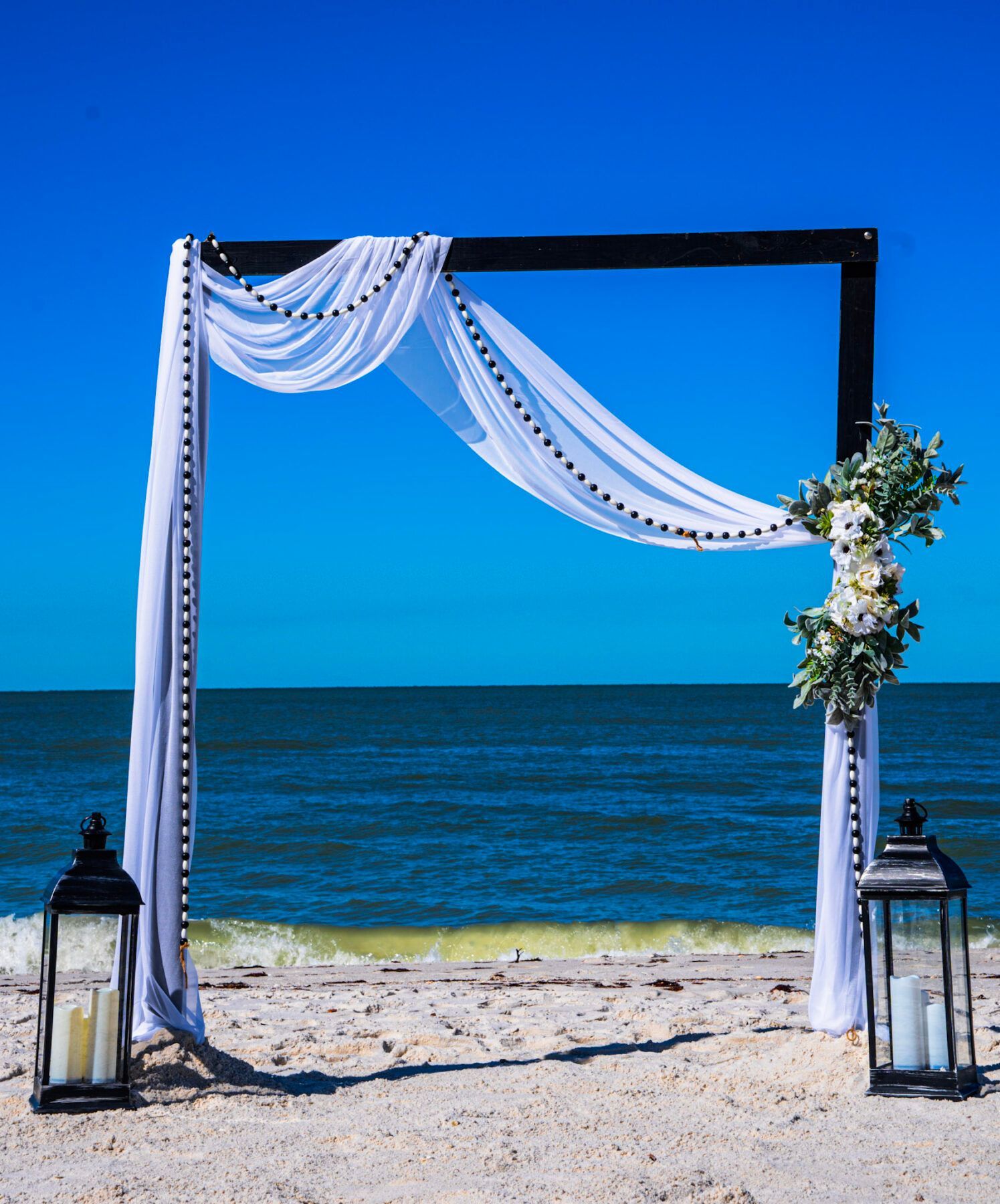 Wedding arch decorated with white fabric and flowers on a beach, blue sky and ocean in background.