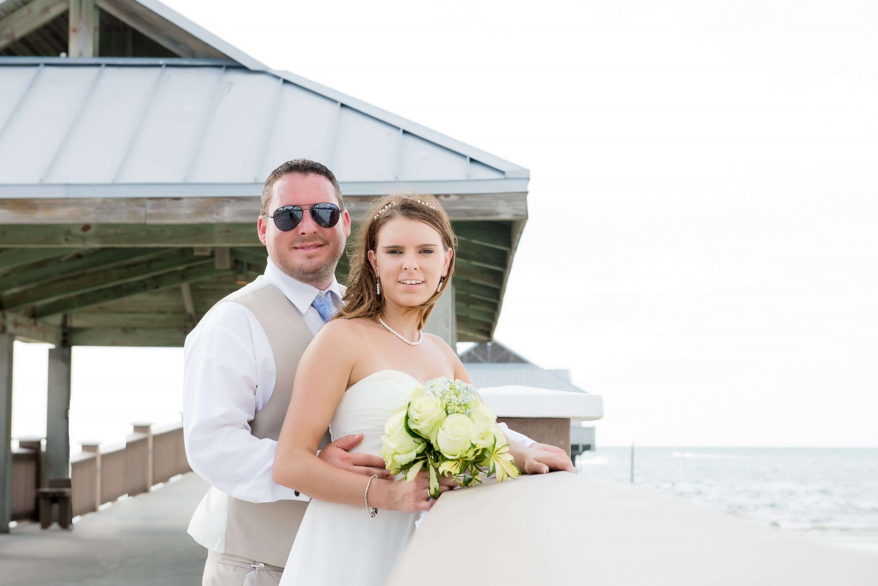 Couple on a pier, bride holding bouquet, groom's arm around her. White dress, tan suit. Ocean in the background.