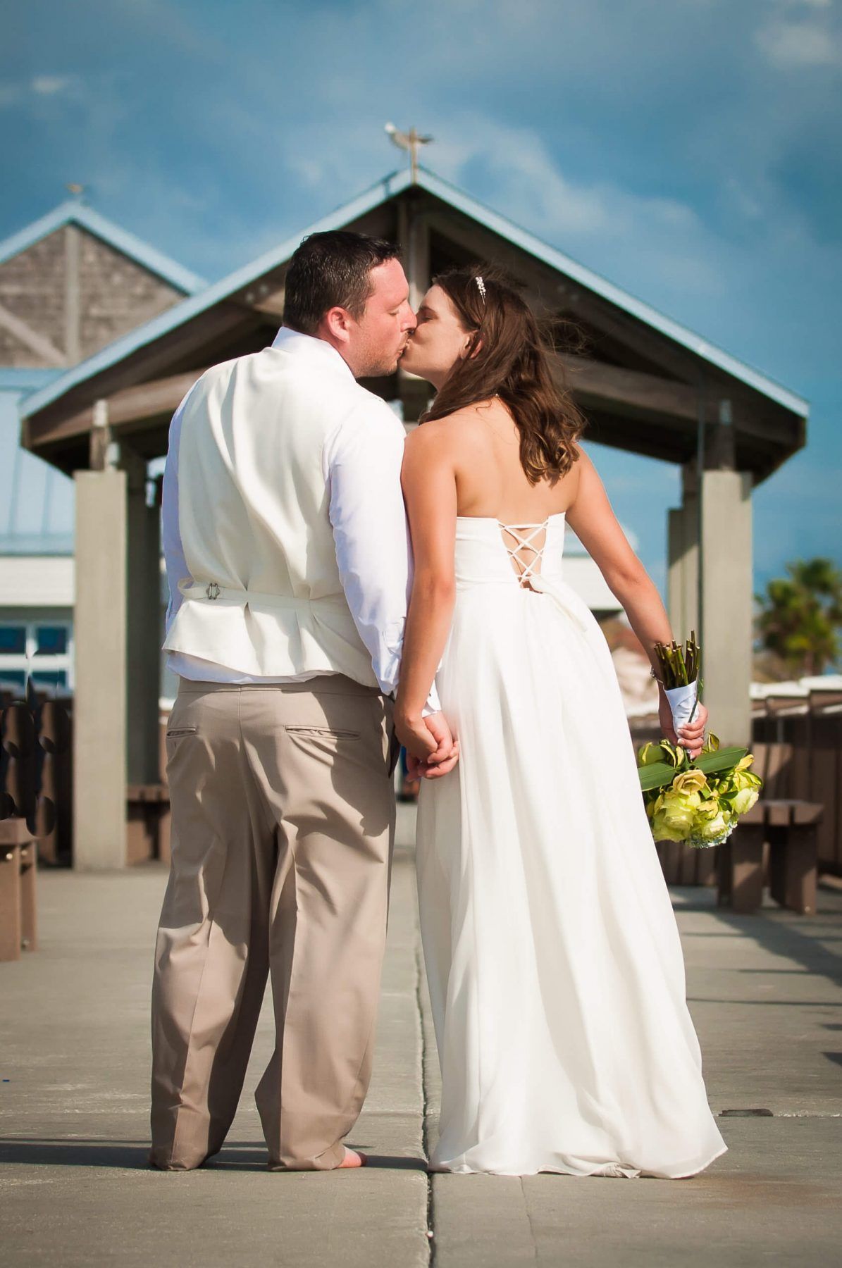 Bride and groom kissing, back to camera, on wooden dock. Bride in white dress, groom in vest, blue sky.