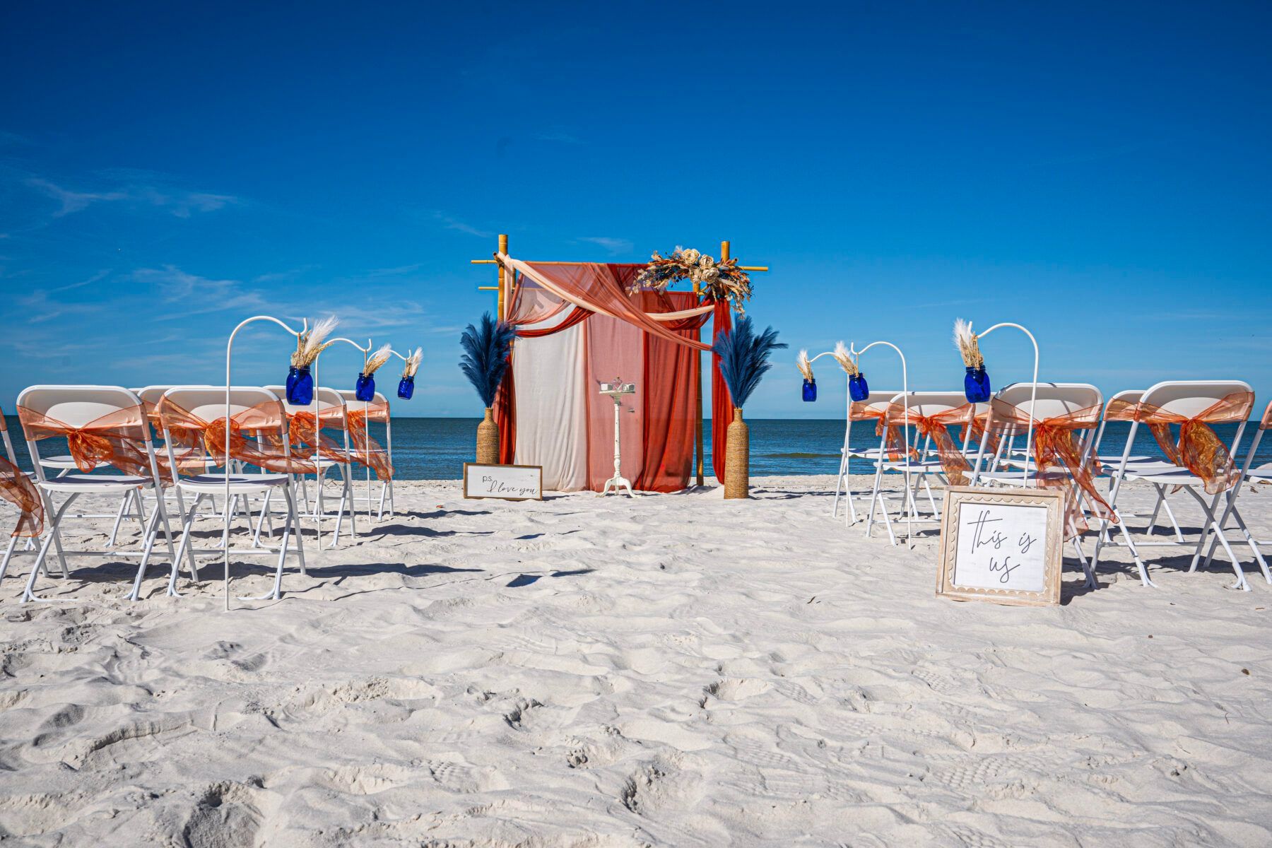 Beach wedding ceremony setup: white chairs with orange accents, arch draped in fabric, blue sky.