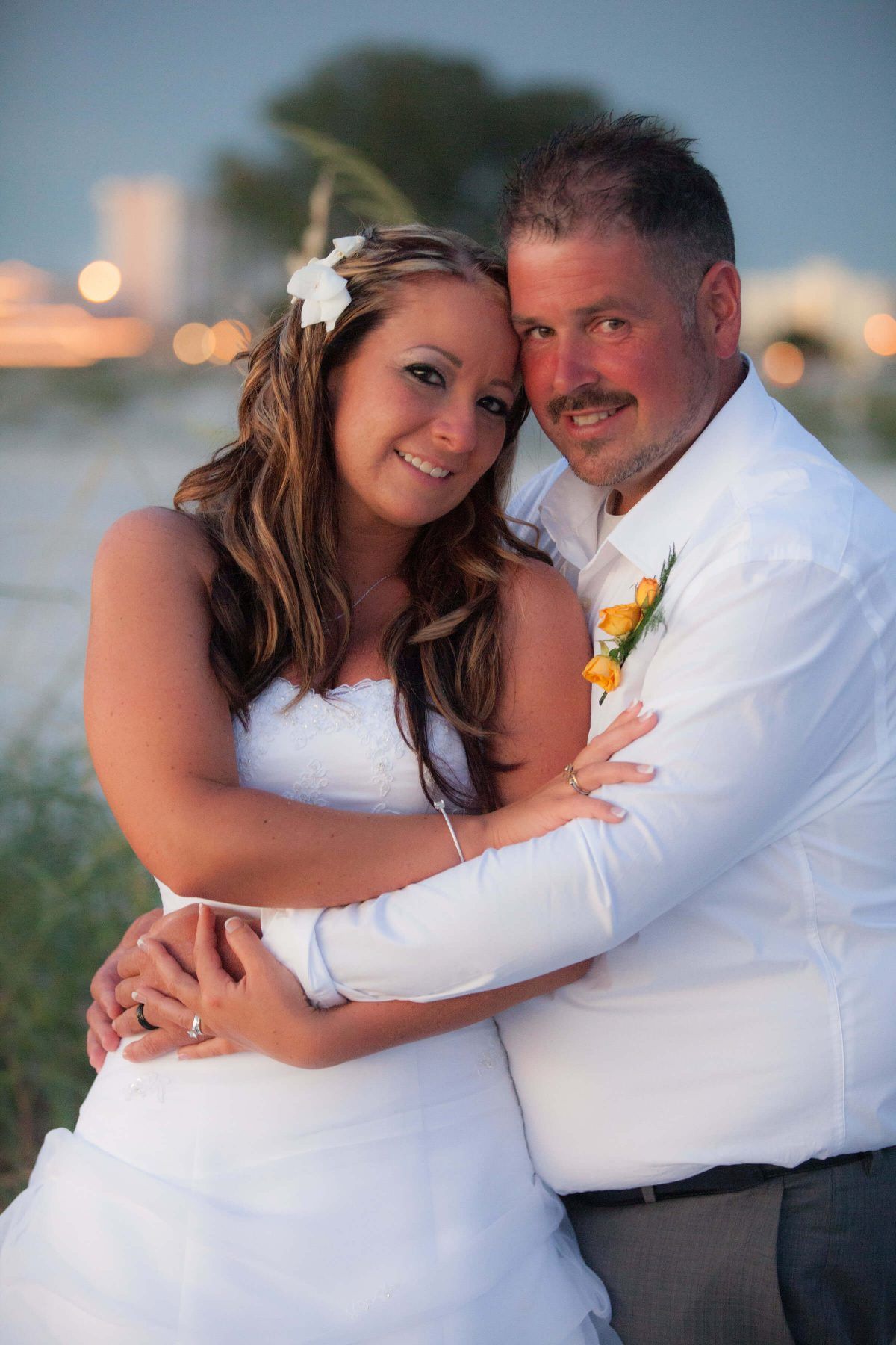 Bride and groom embrace on a beach at dusk. She wears a white gown and flower in her hair; he wears a white shirt.