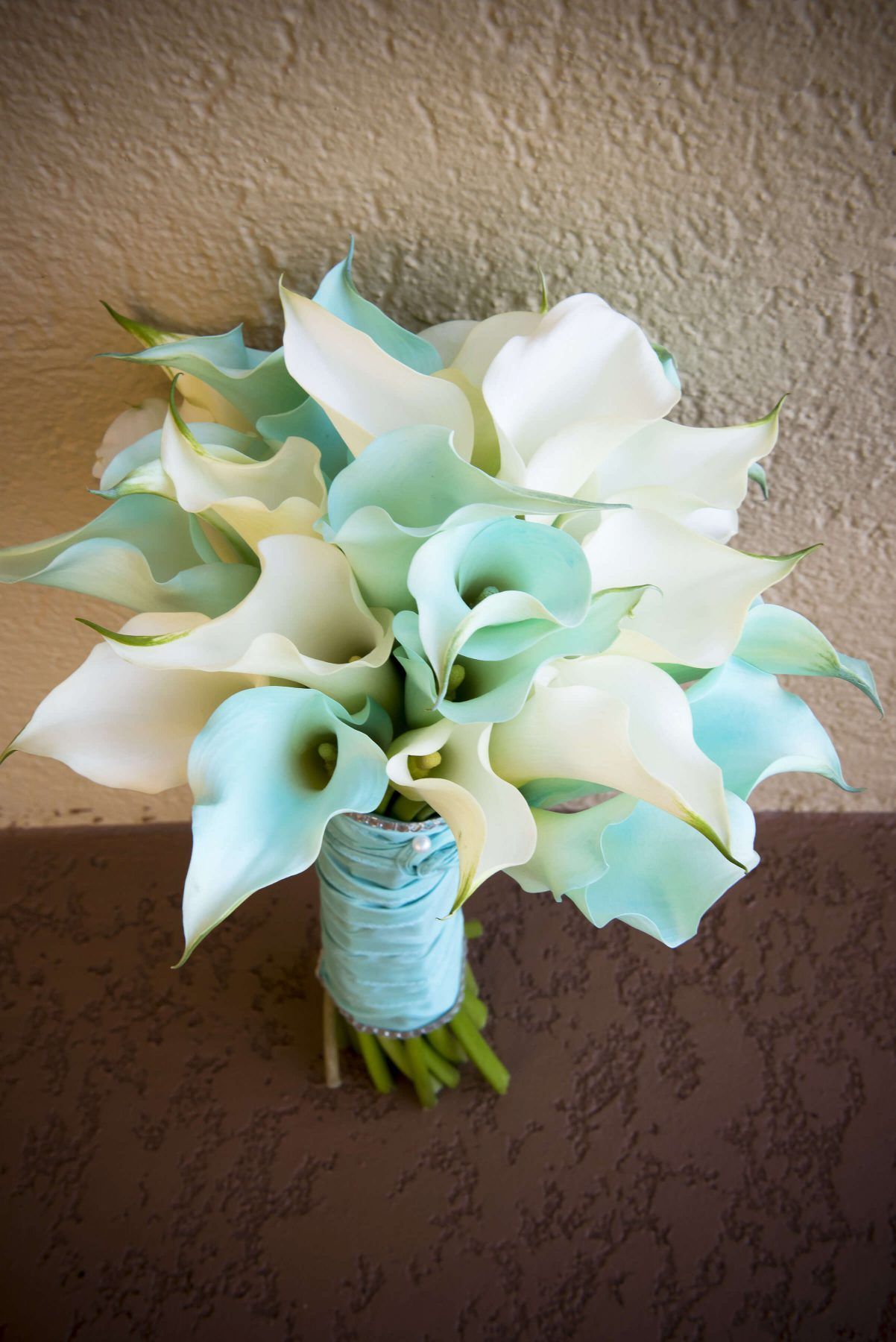 Bouquet of white and teal calla lilies, stems wrapped in light blue ribbon, against a brown and tan wall.