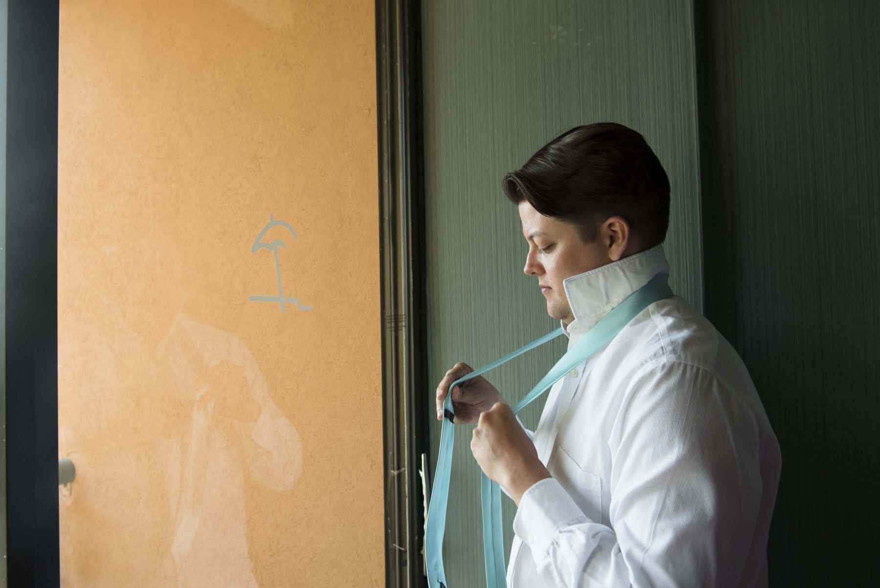 Person in white shirt and blue tie, tying the tie, standing near a window.