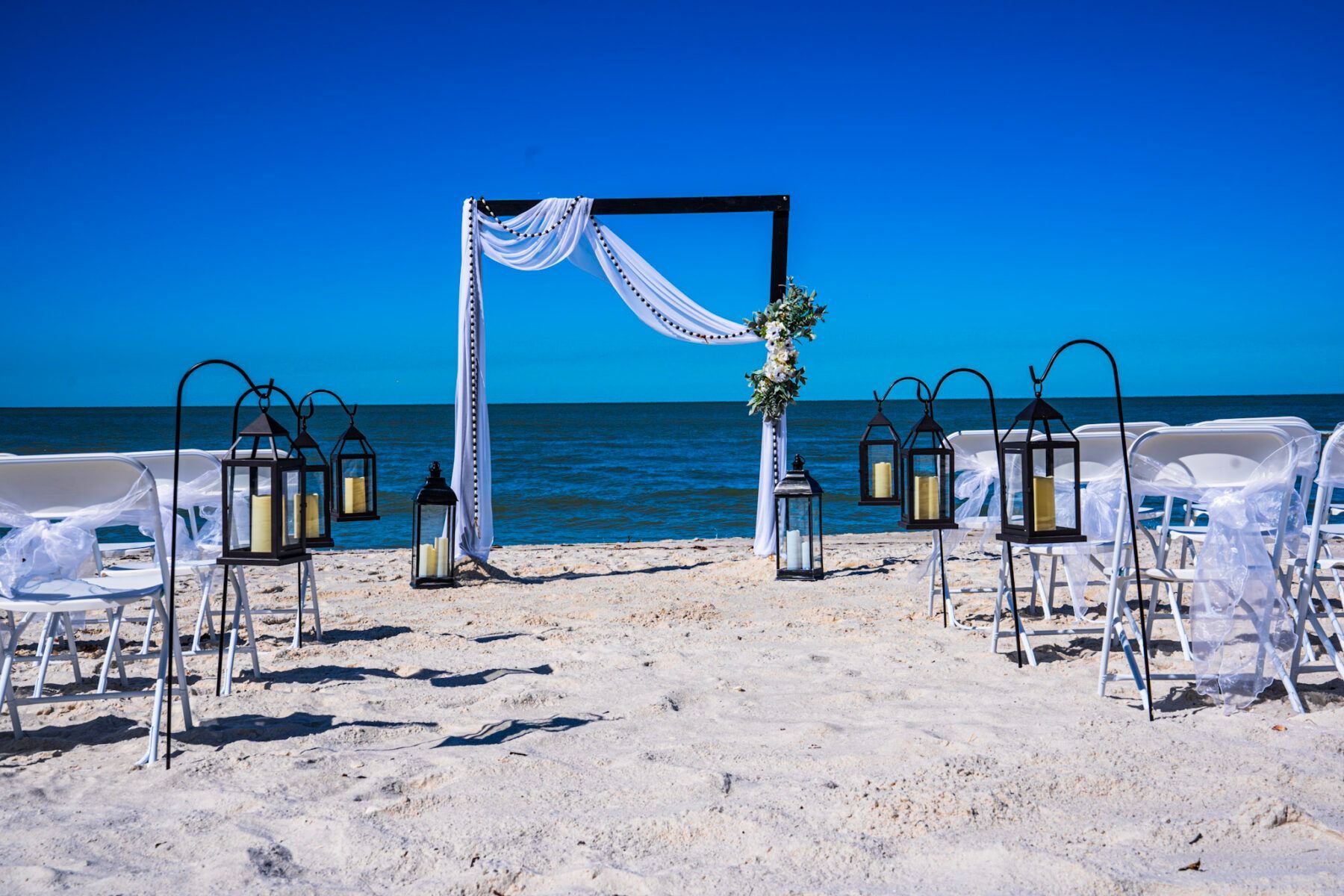 Beach wedding setup with arch, lanterns, chairs, and ocean view.