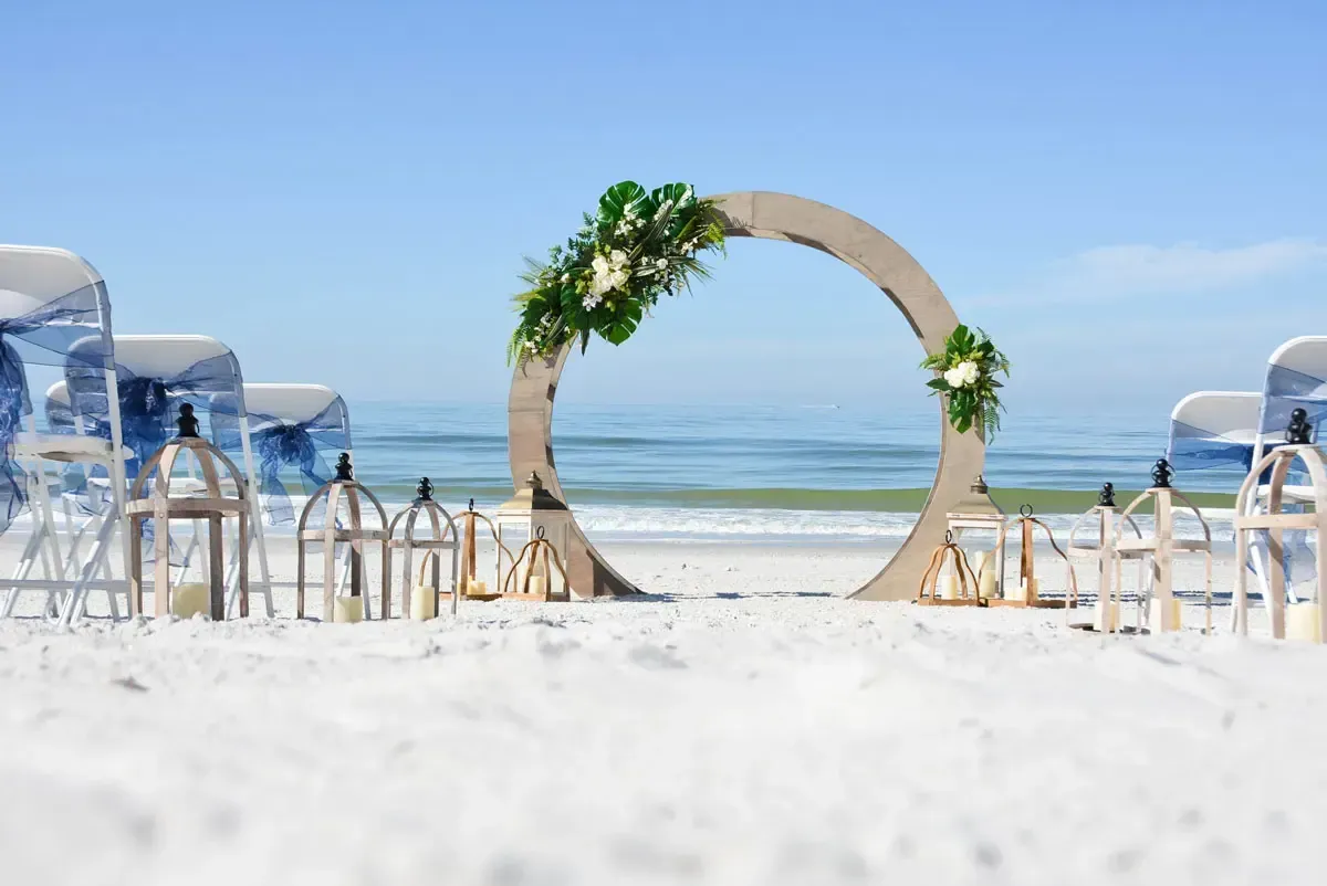 Wedding ceremony setup on a sandy beach with a floral arch, blue chairs, and lanterns, overlooking the ocean.