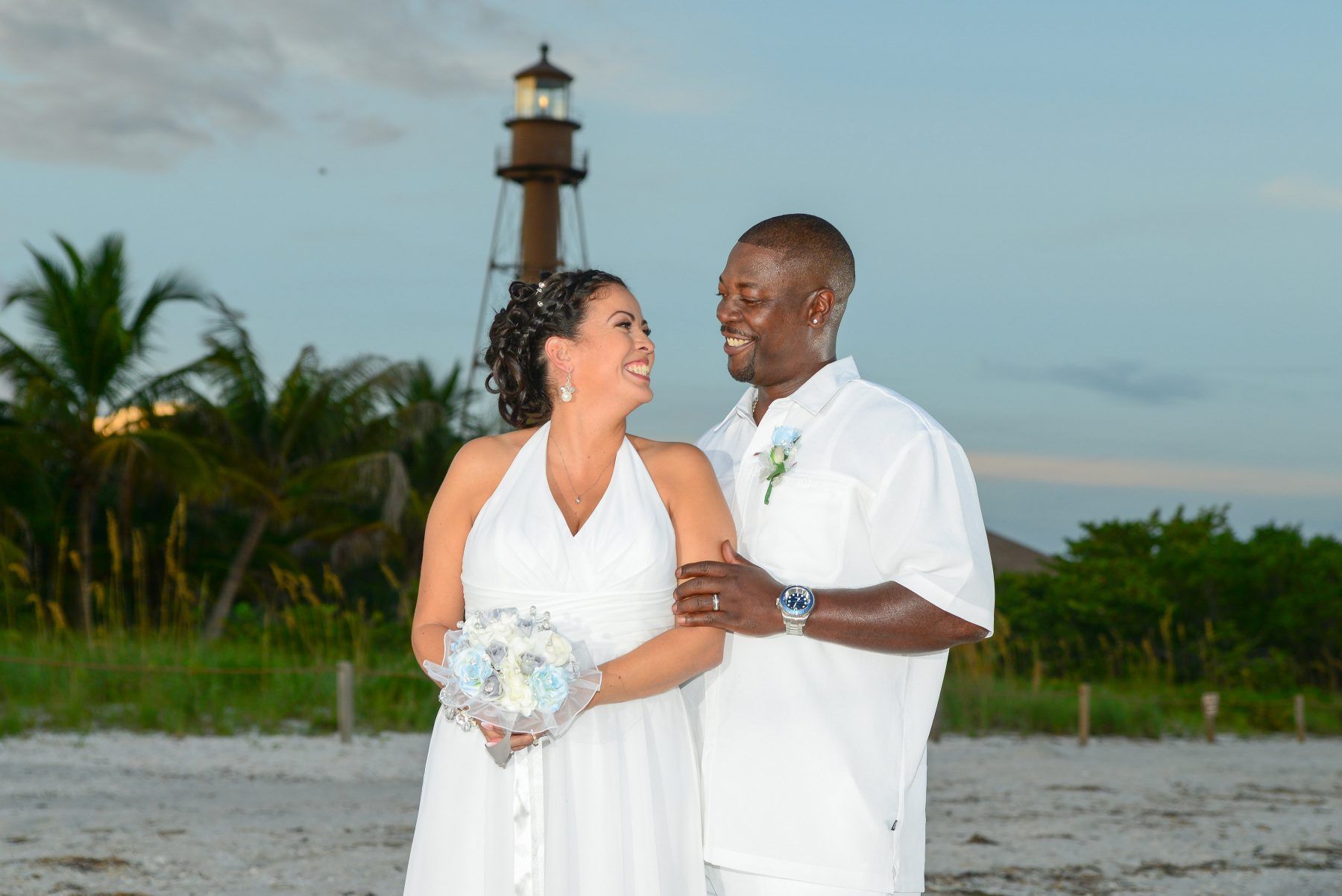 Couple smiles at each other on a beach, near a lighthouse. Bride in white dress, groom in white shirt.