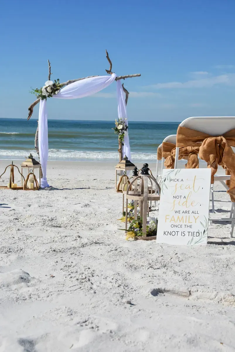 Beach wedding setup: arch with white fabric, chairs with gold bows, lanterns, and a sign on a sunny day.