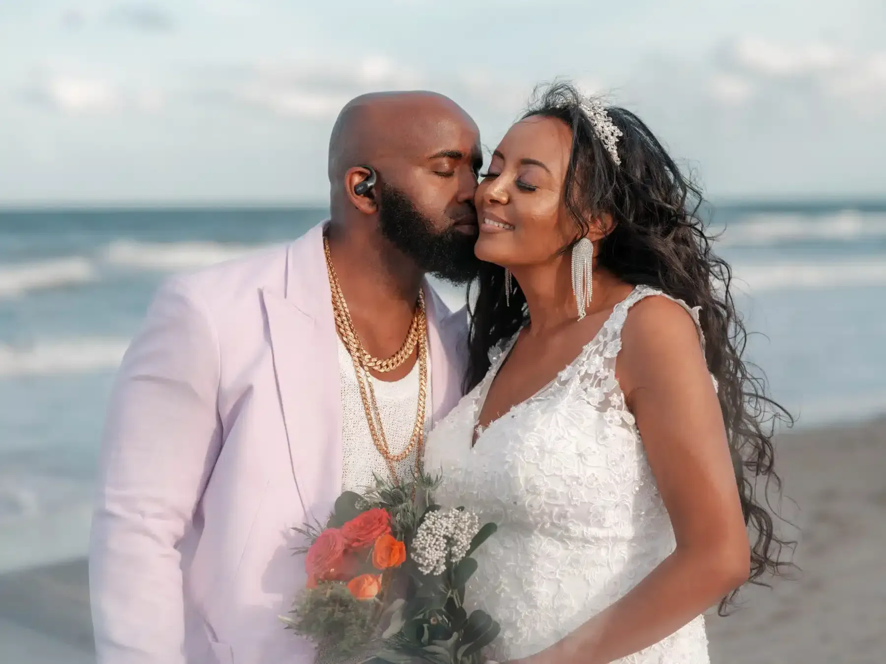 Couple kissing on a beach; man in lavender jacket, woman in wedding dress. Ocean in background.