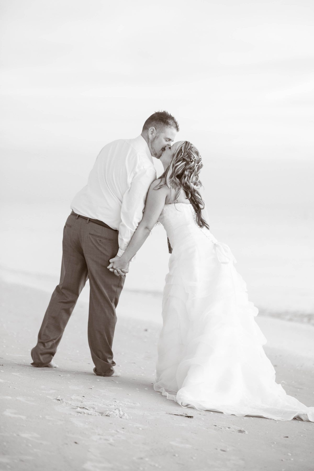Couple kissing on a beach. The woman is wearing a wedding dress and the man is in dress pants and a shirt.