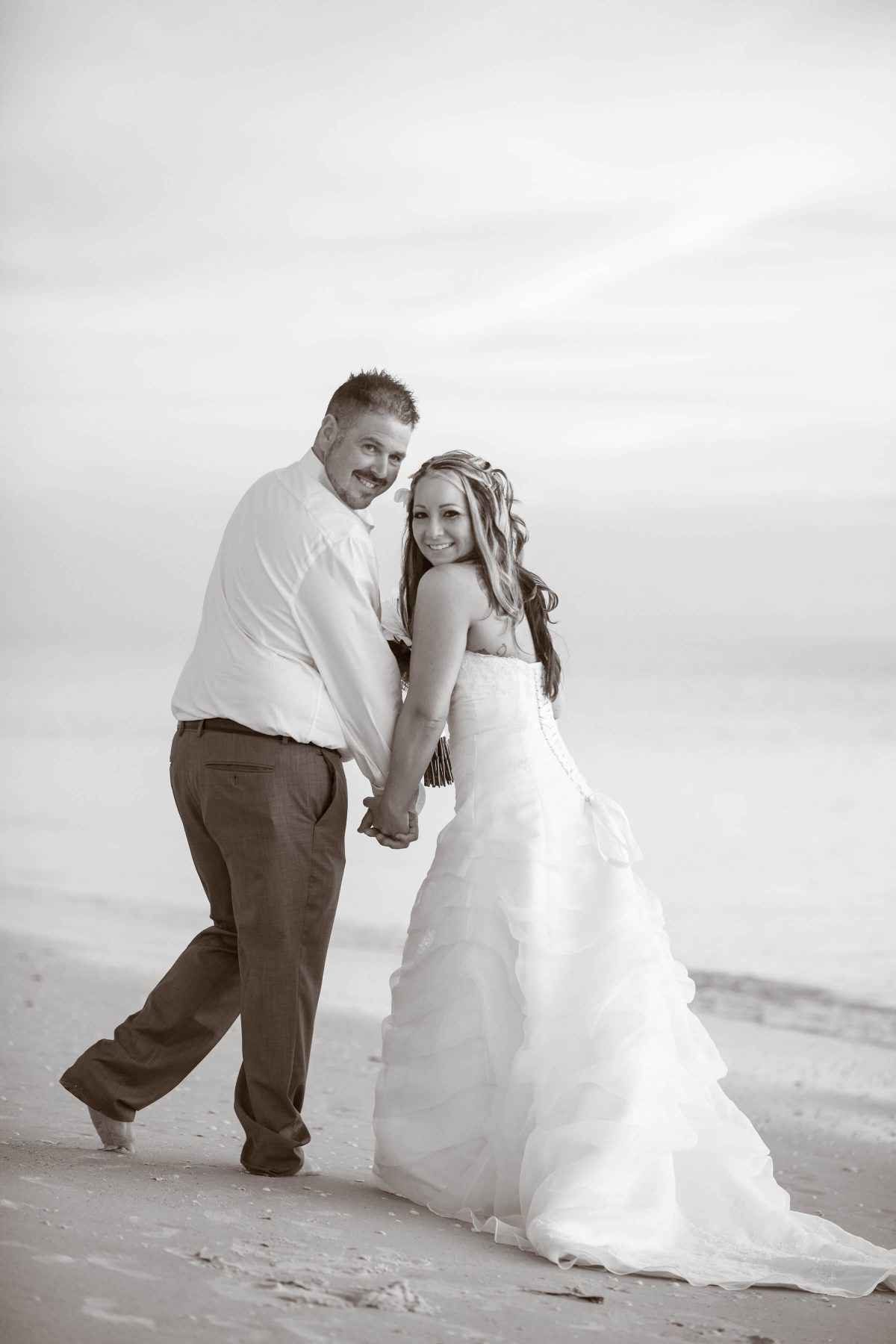 Bride and groom on a beach, holding hands and smiling. She wears a white gown, he wears a shirt and pants.