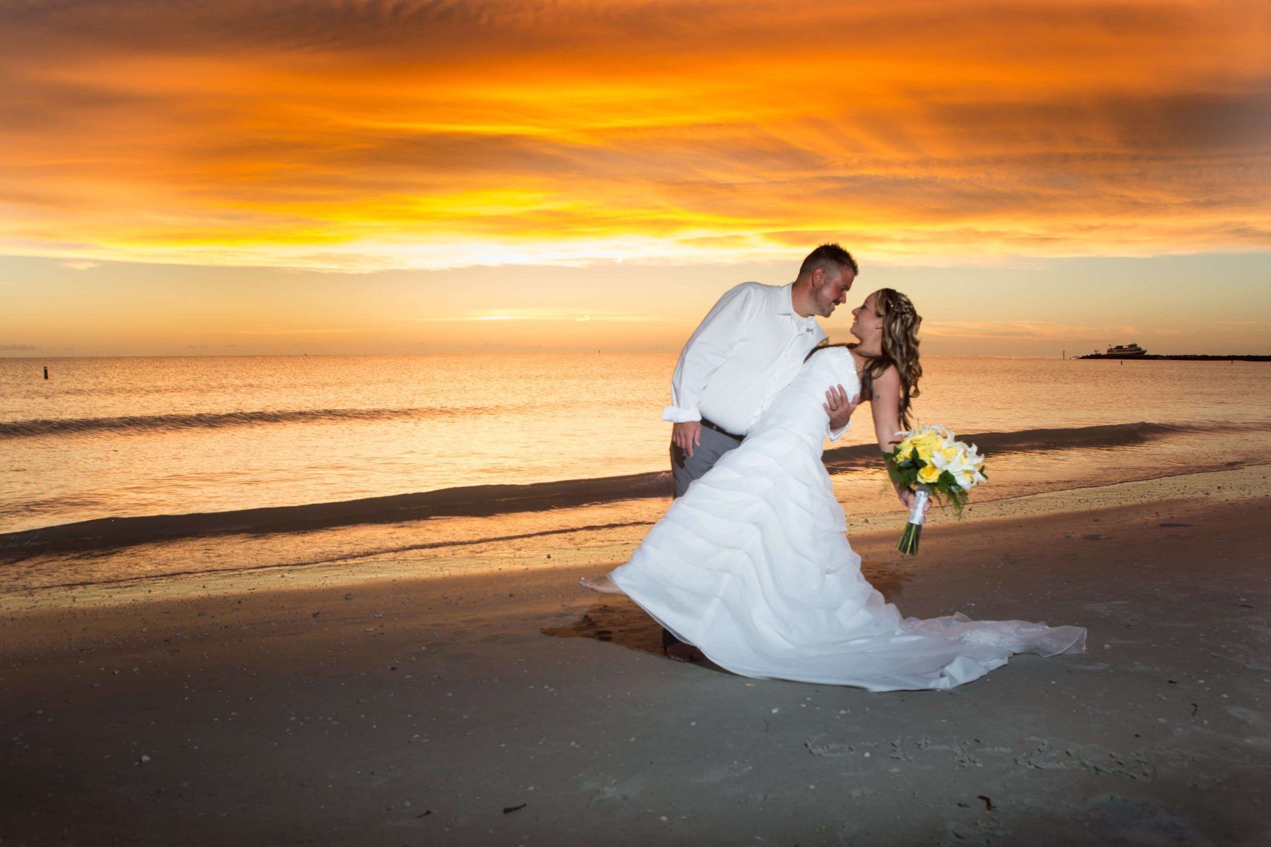 Couple embraces on beach at sunset; woman in wedding dress, holding flowers, man in white shirt.