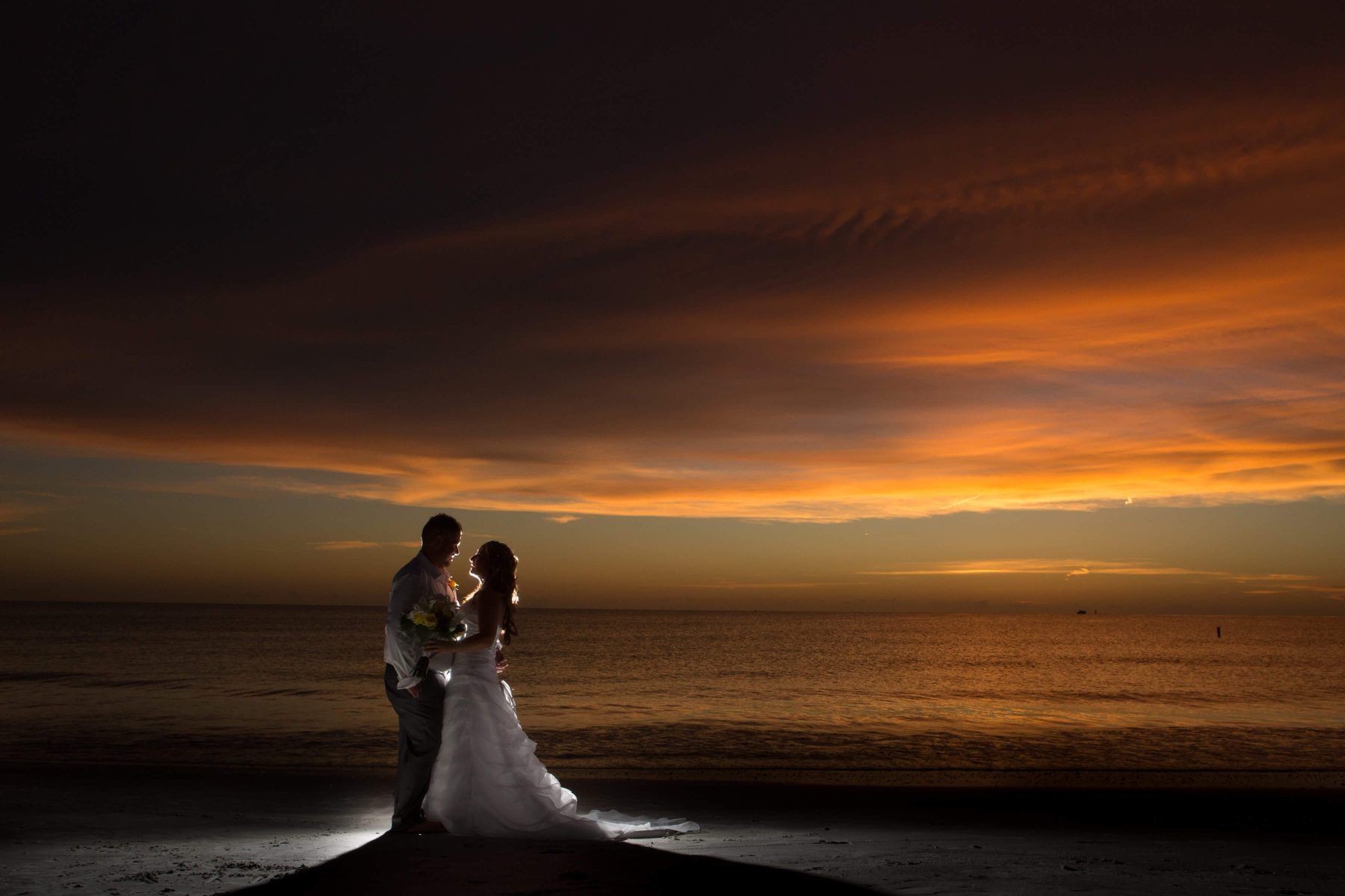 Couple on beach at sunset, silhouetted against orange sky, bride in white dress.