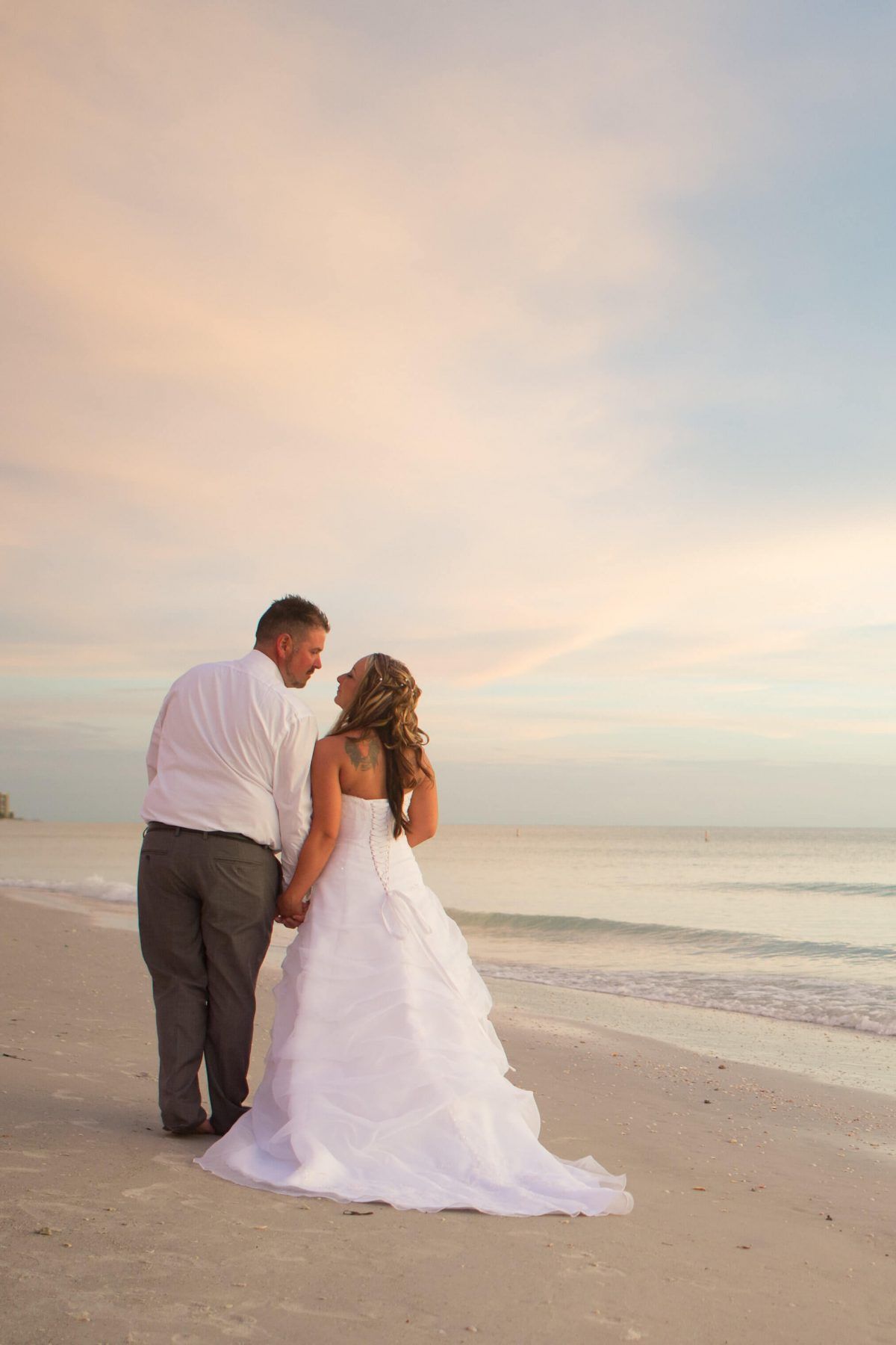 Newlyweds walking hand-in-hand on a beach at sunset. Bride in white dress, groom in shirt and pants.