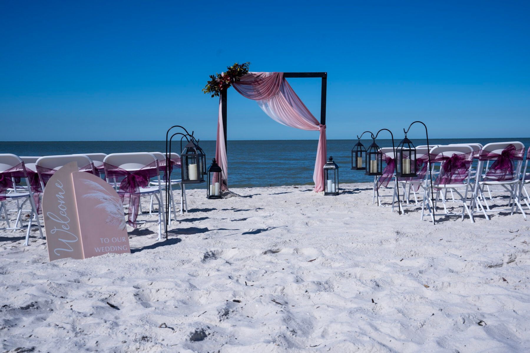 Beach wedding setup with chairs, arch, and lanterns on white sand under a blue sky.