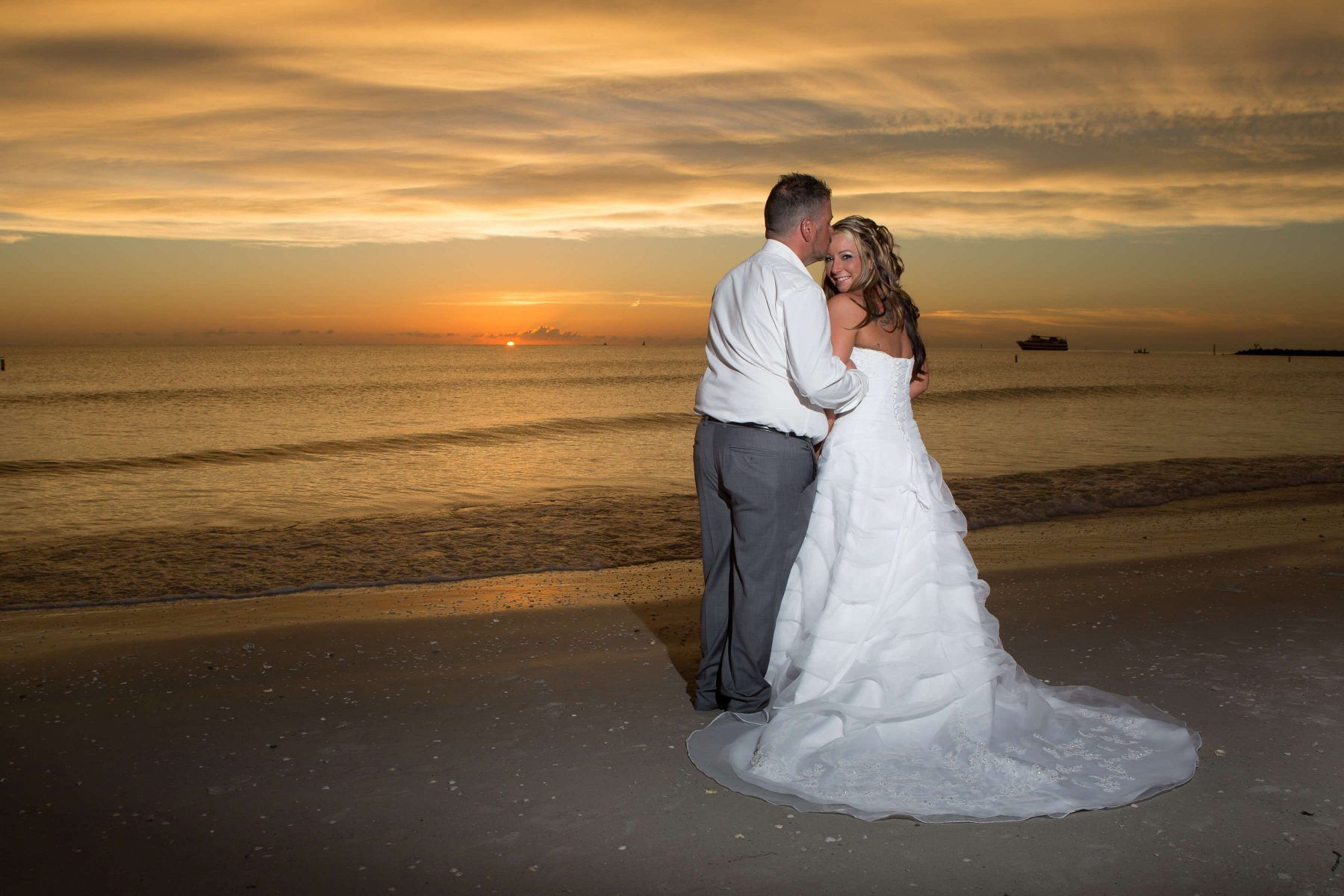 Bride and groom embrace on a beach at sunset. The bride wears a white dress, the groom a white shirt and gray pants.