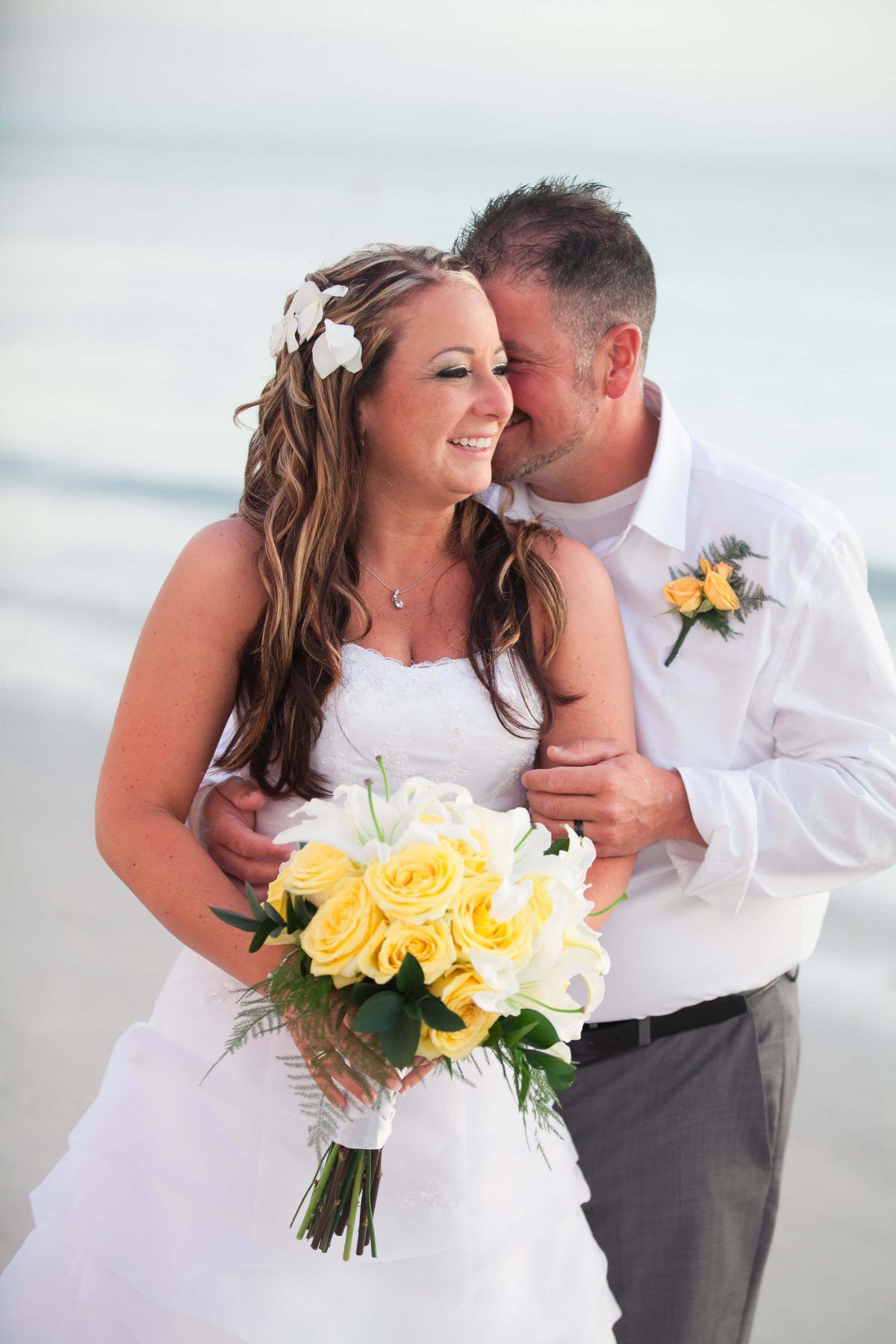 Bride and groom smiling on a beach. Bride holds yellow roses, groom embraces her; ocean in background.