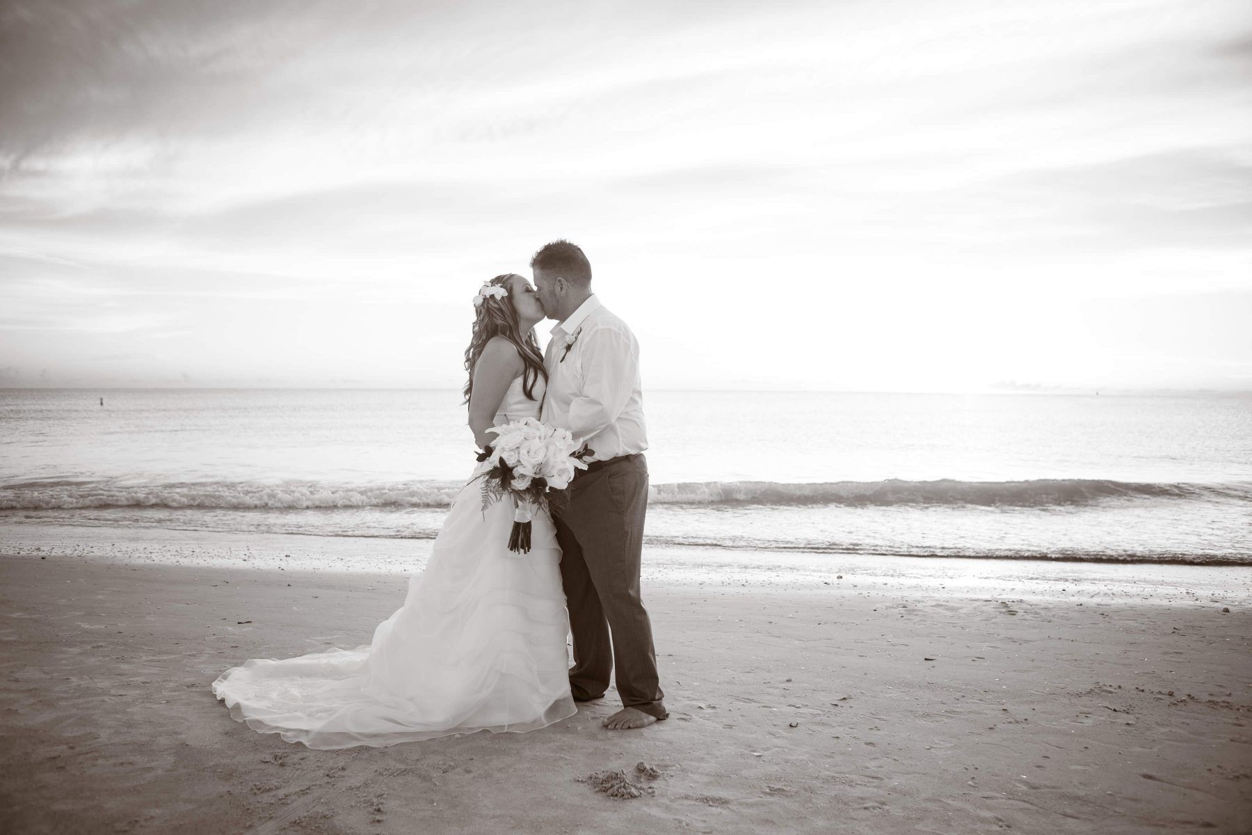 Couple kissing on a beach at sunset; woman in wedding dress, man in shirt and pants.