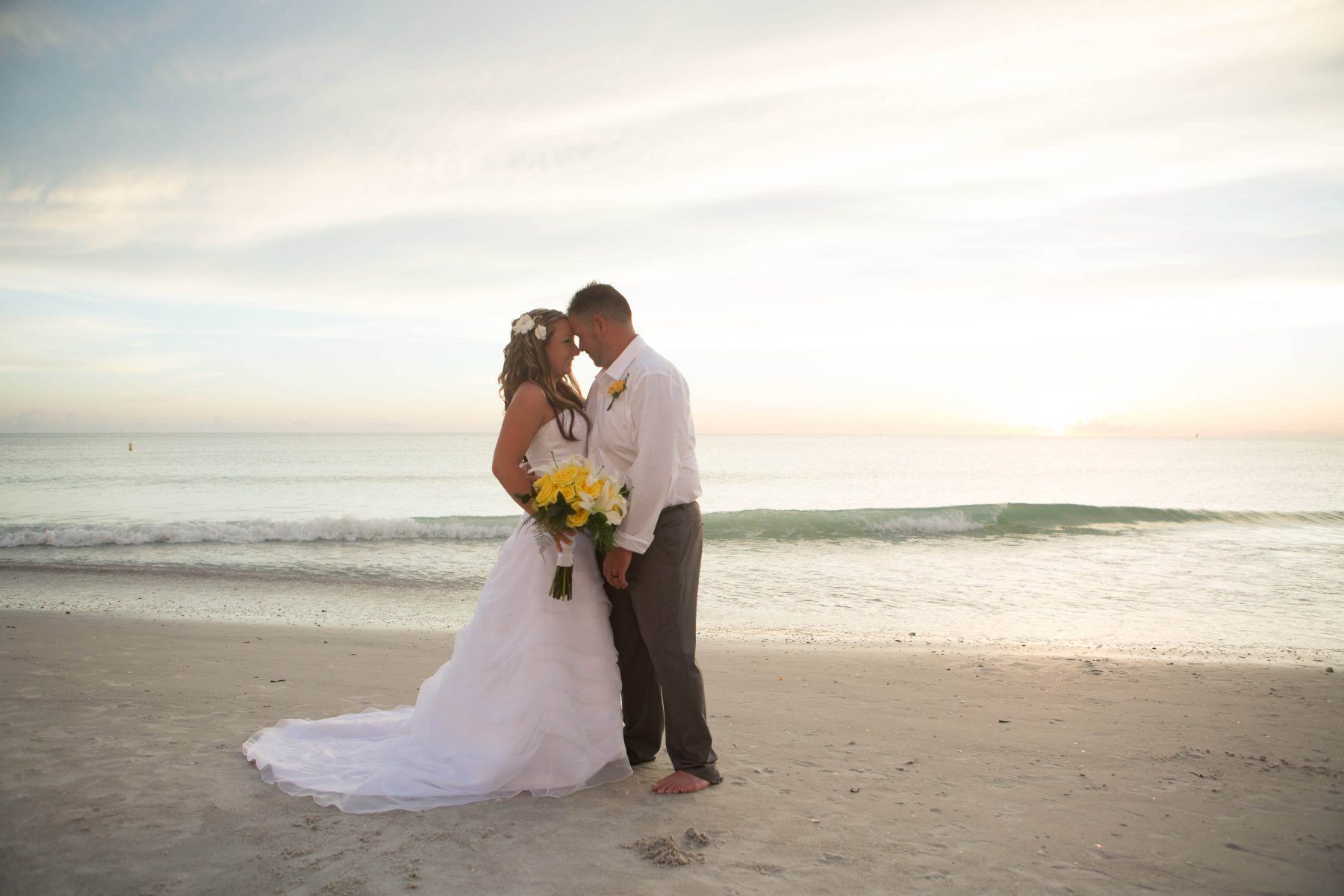 Couple embraces on a beach at sunset; bride in white gown, groom in dress shirt, holding yellow bouquet.