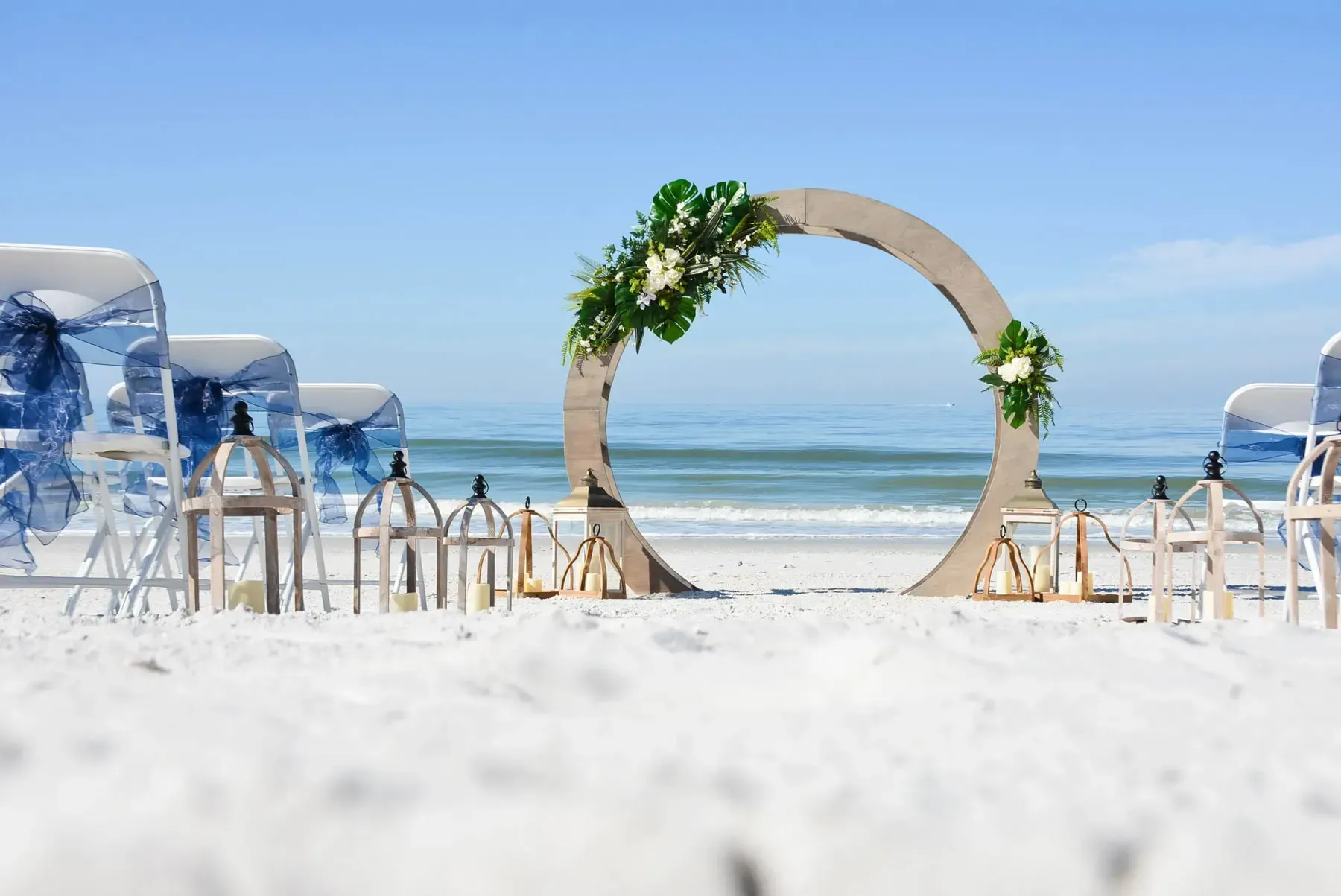 Wedding ceremony setup on a sandy beach. A circular arch is decorated with flowers, ocean in the background.