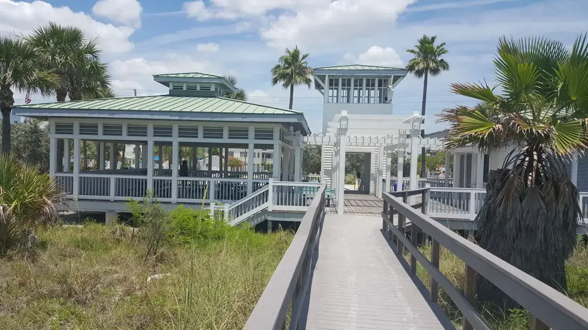 Wooden boardwalk leading to white gazebo-like structures, with palm trees and a blue sky.