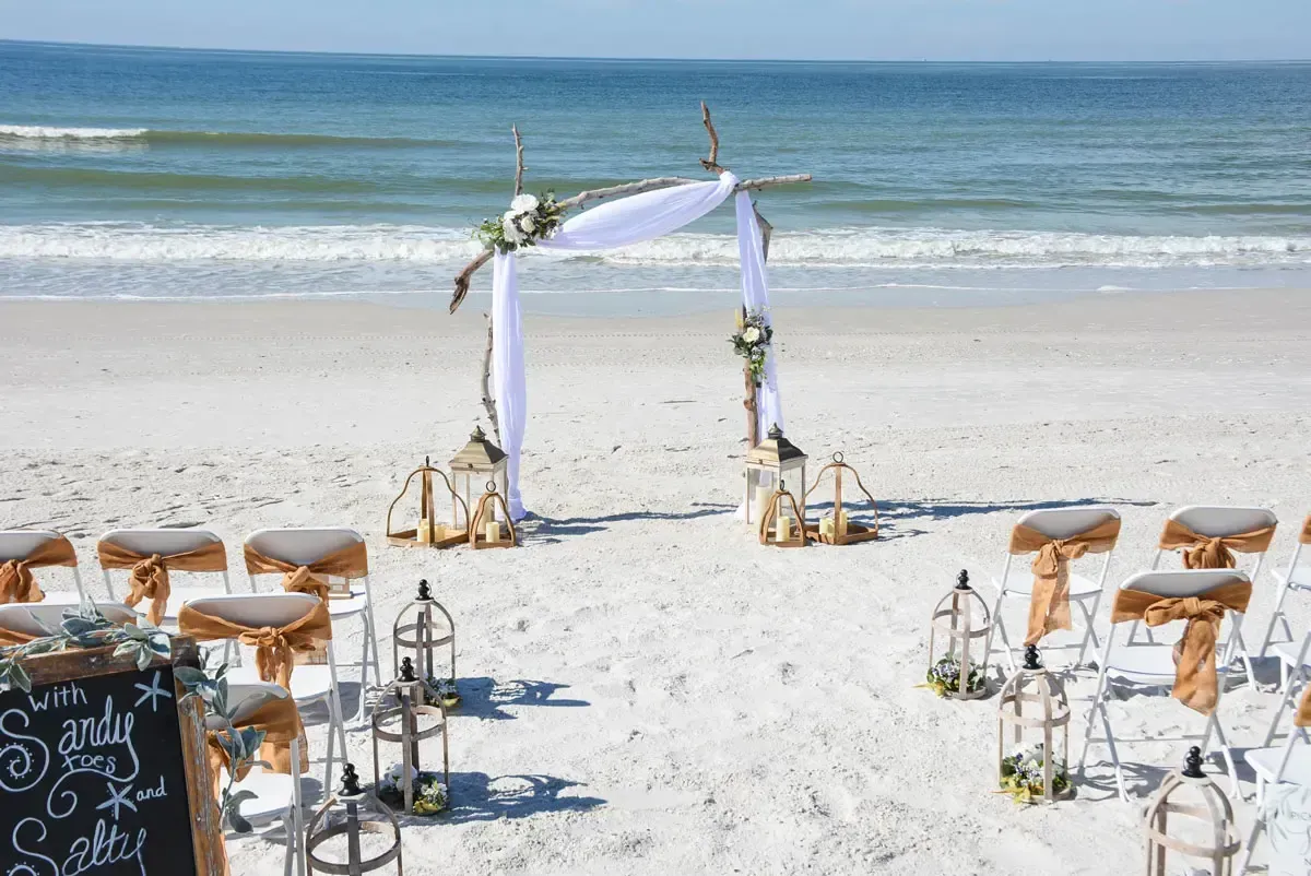 Beach wedding setup with arch, chairs, and lanterns. Ocean in background.
