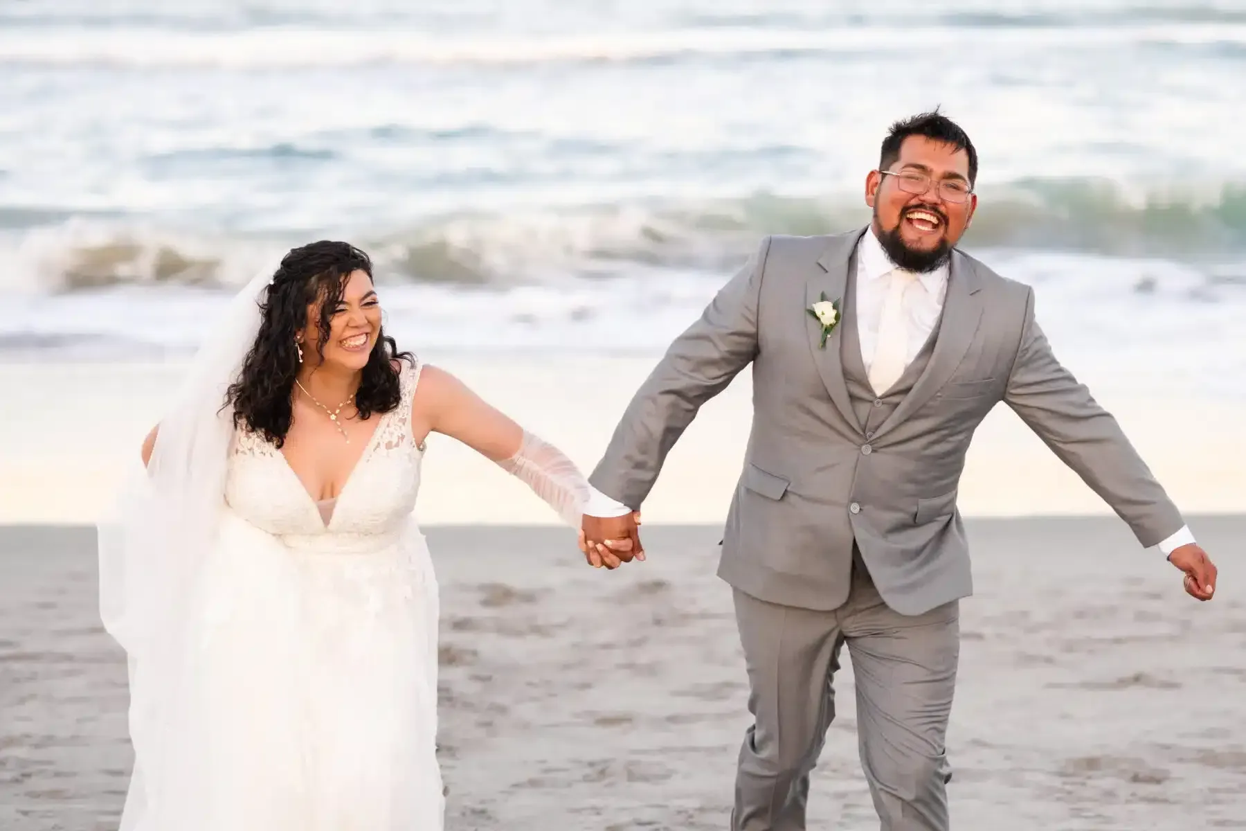 Couple holding hands, laughing, running on a beach, waves in background. Bride in white dress, groom in gray suit.