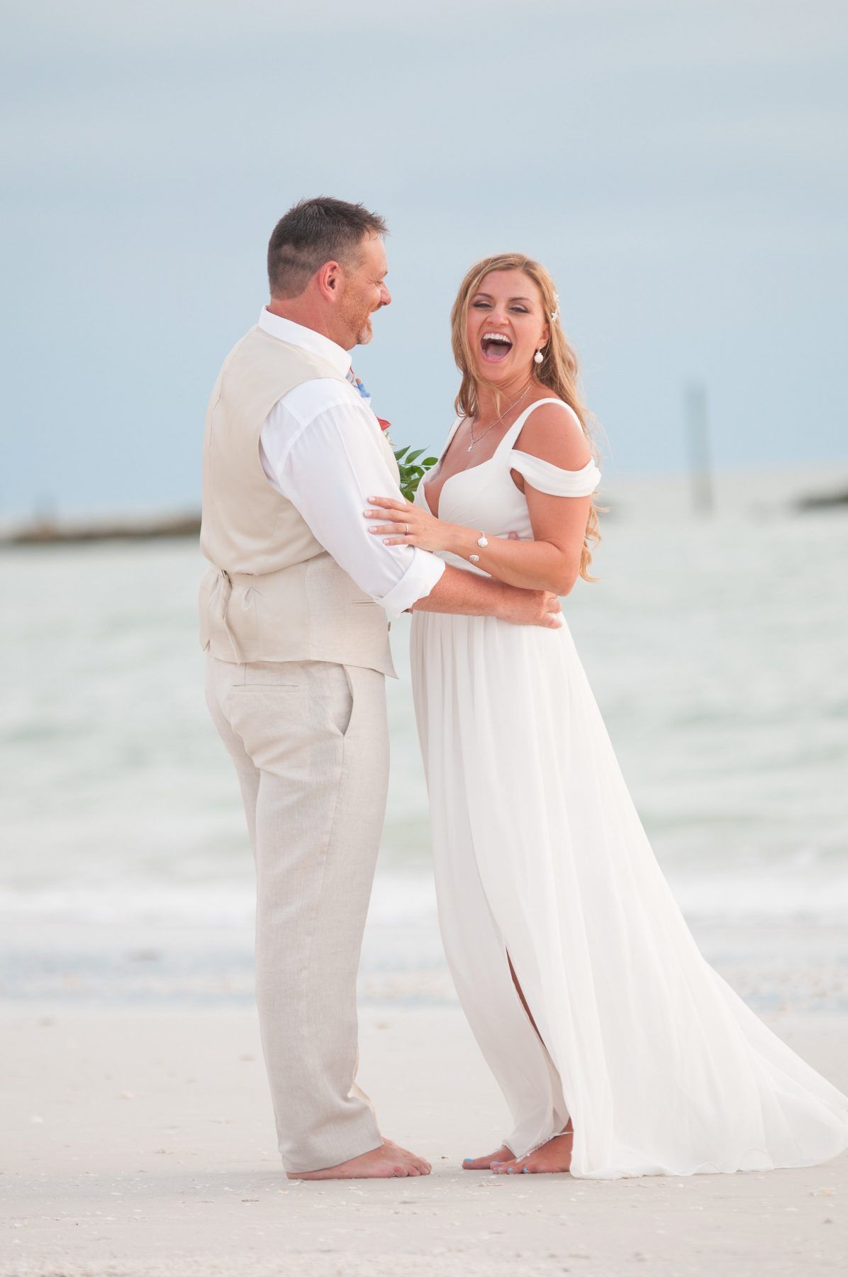 Bride and groom laughing on a beach, both barefoot. The bride in white gown, the groom in tan vest. Ocean in the background.