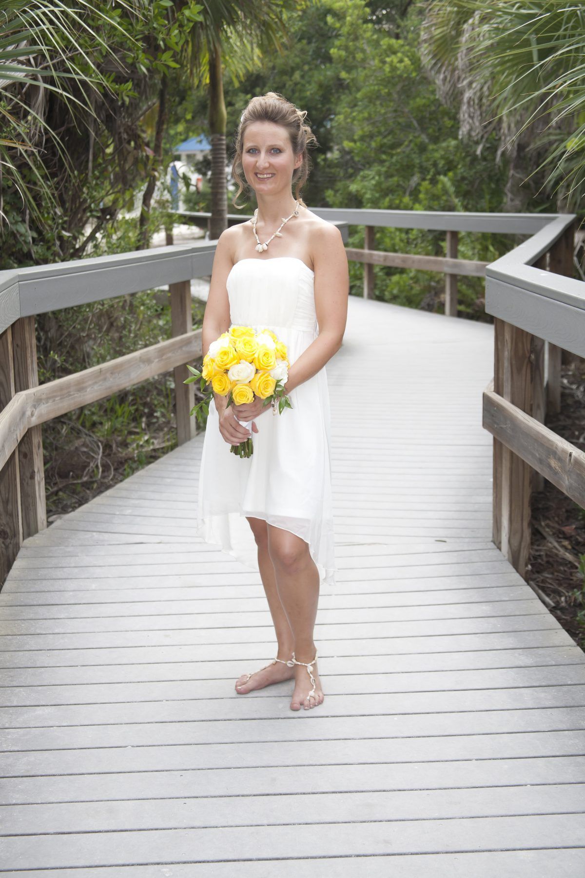 Woman in white dress holds yellow flowers on wooden walkway.