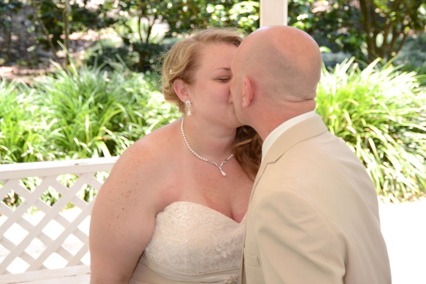 Bride and groom share a kiss, under a white gazebo with greenery background.