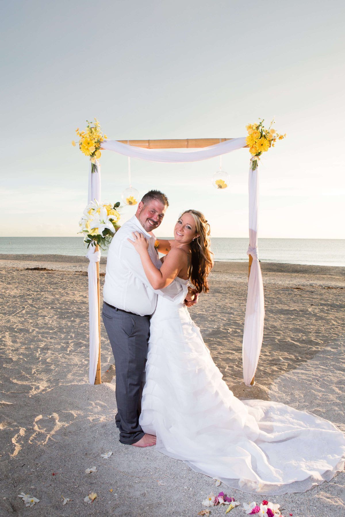 Couple embraces on a beach in front of a wedding arch decorated with yellow flowers; ocean in the background.