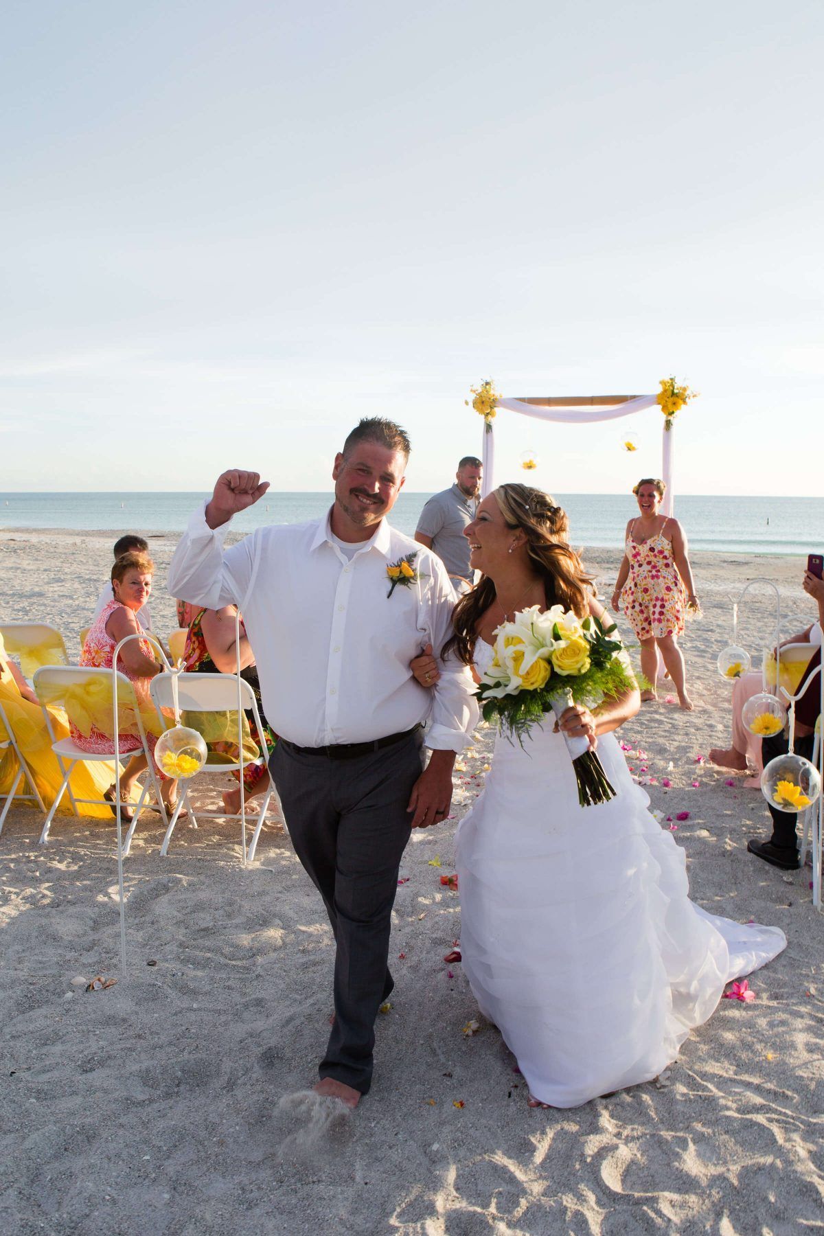 Bride and man exiting beach wedding. Bride laughs, holding flowers. Man pumps fist. Yellow décor.