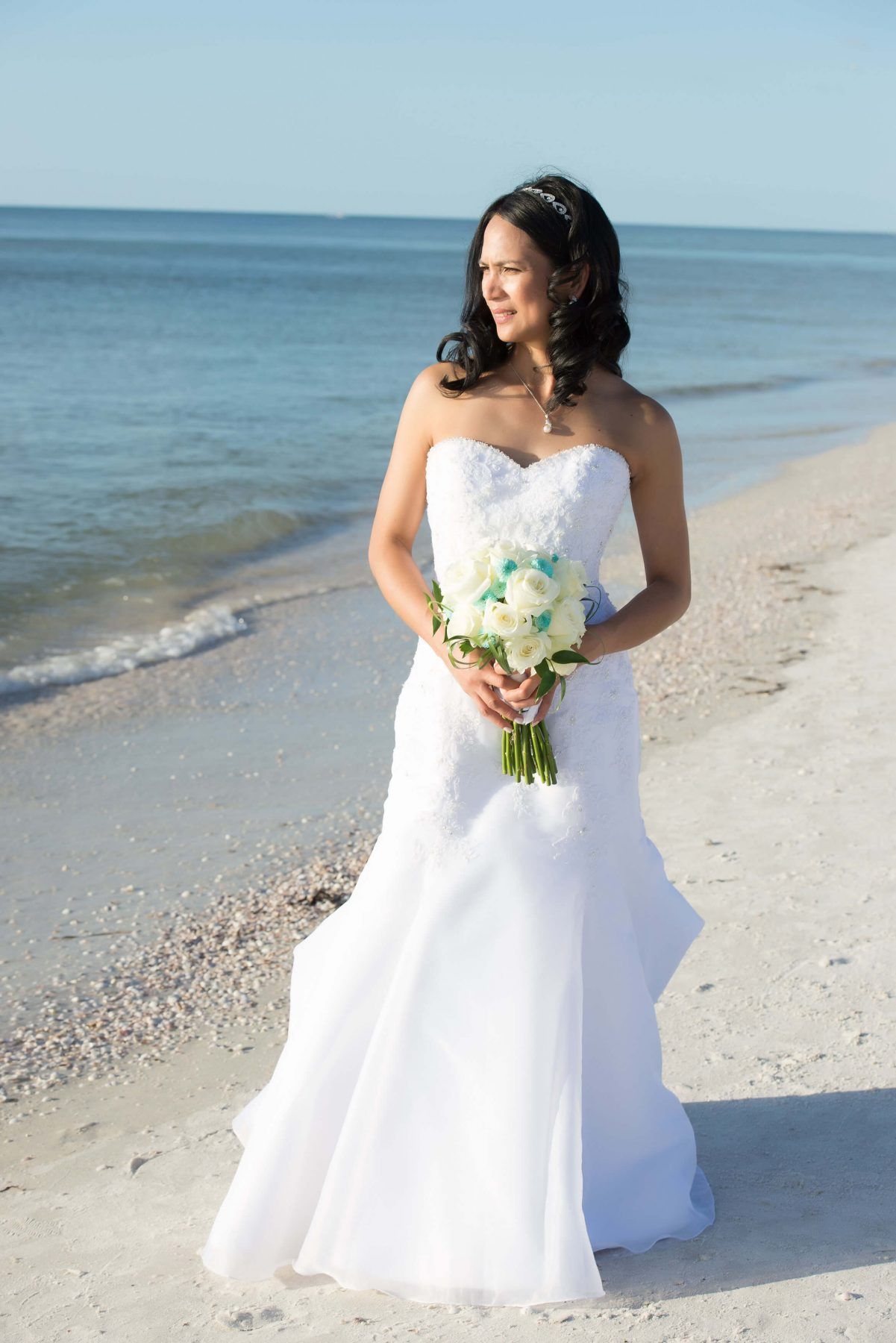 Bride in white strapless gown holds bouquet, stands on beach, looking to the side. Ocean in background.