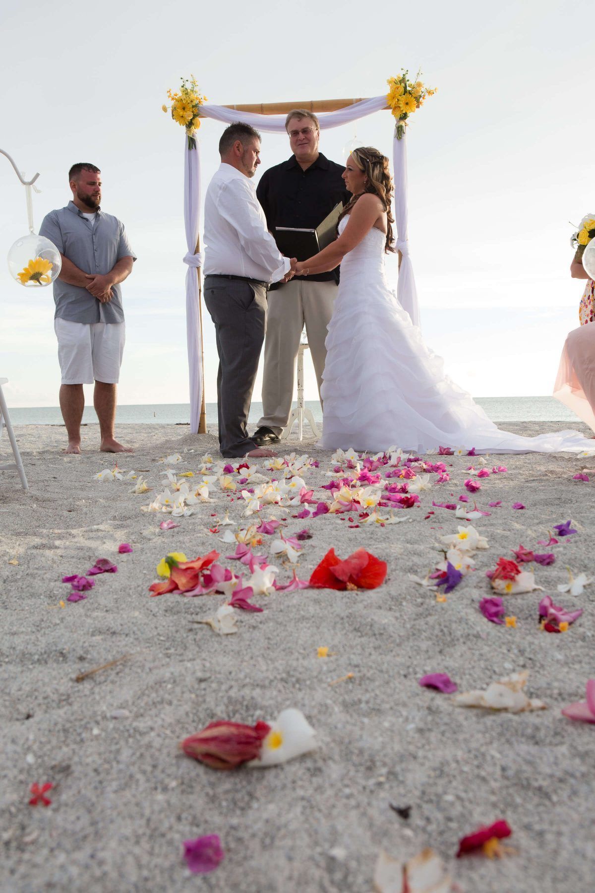 Wedding ceremony on beach; couple holding hands under arch with floral decorations and petal-covered sand.