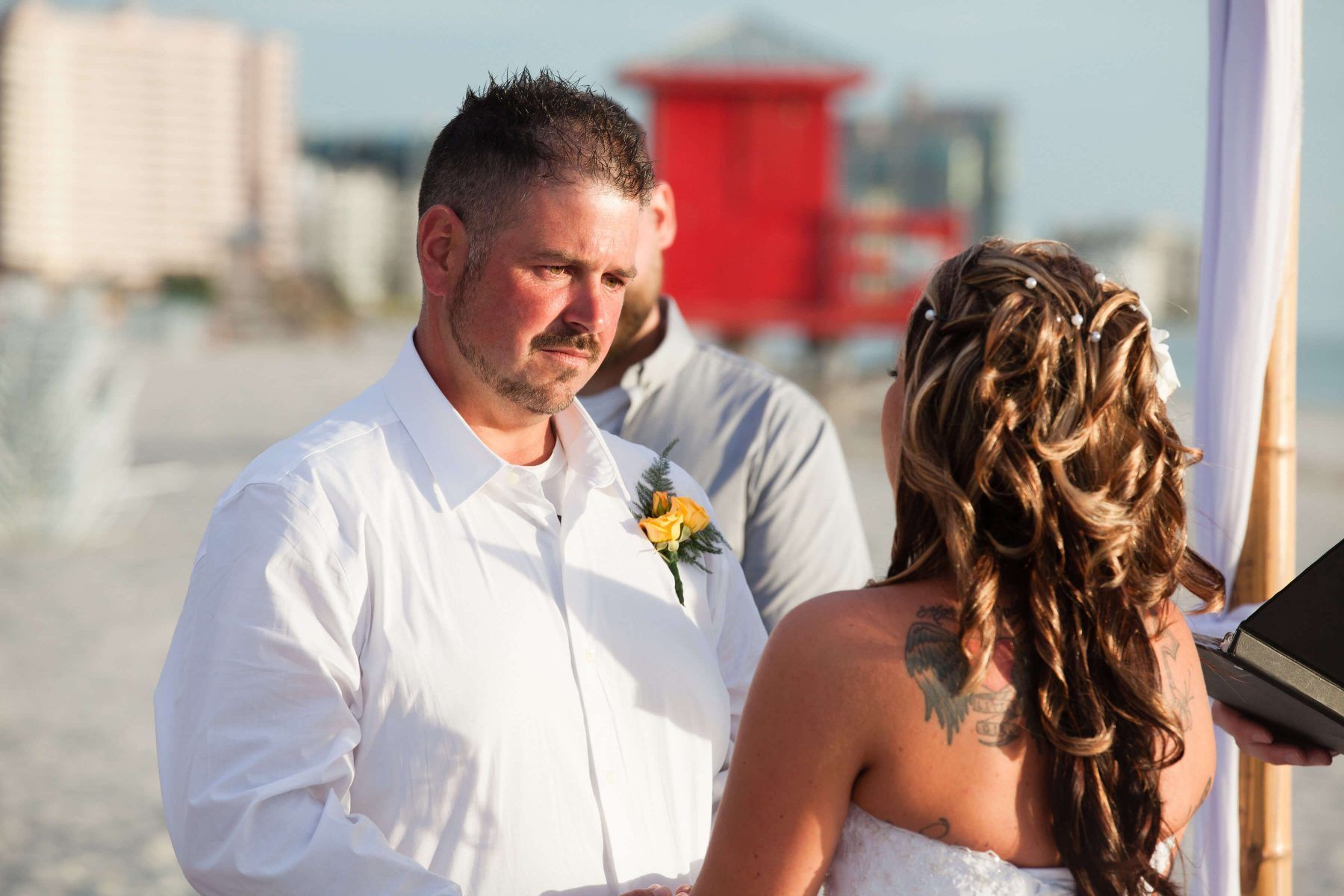Man and woman at a beach wedding ceremony, both facing each other. The man is wearing a white shirt and looking solemn.