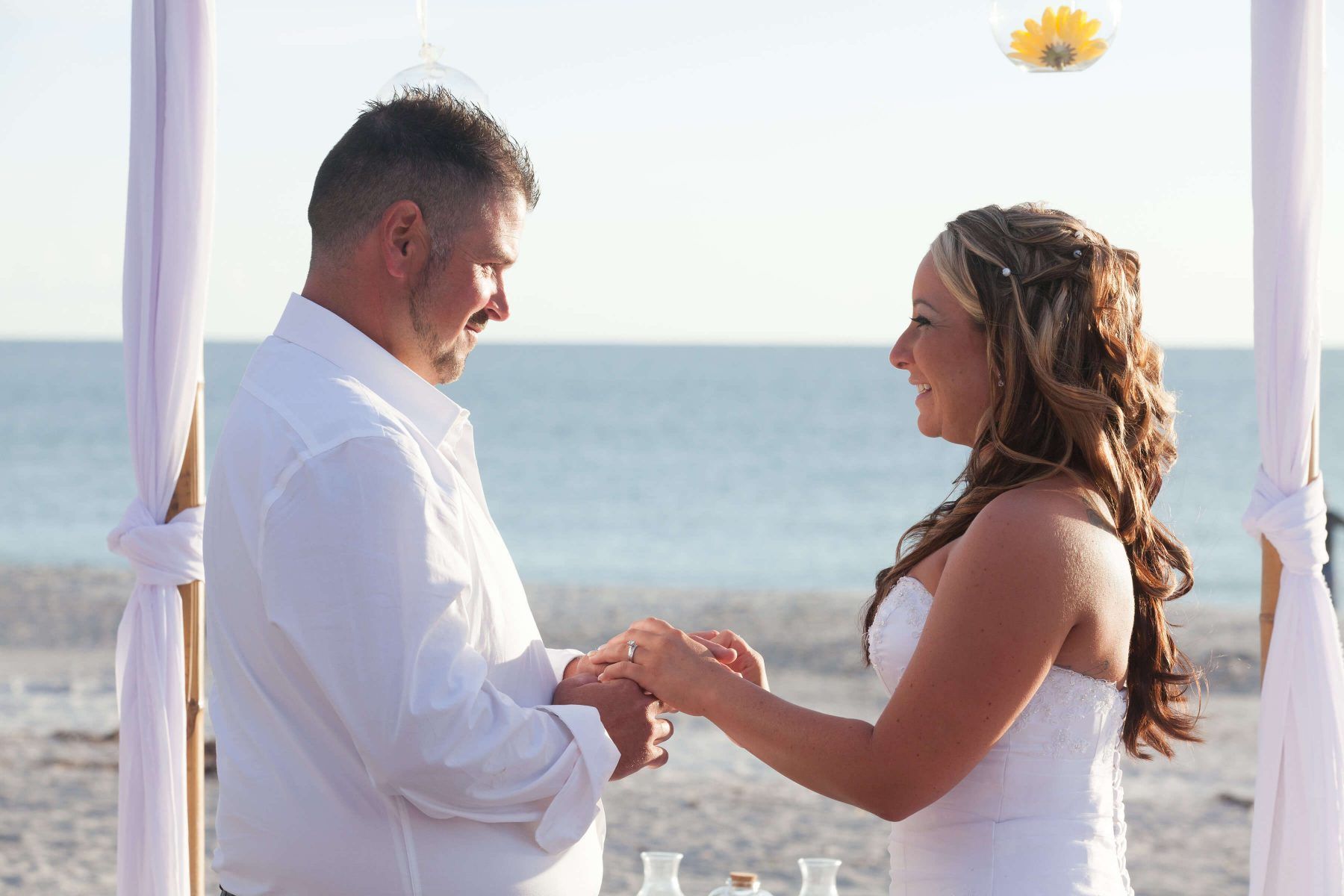 Bride and groom exchanging rings on a beach, under a draped archway, ocean in the background.
