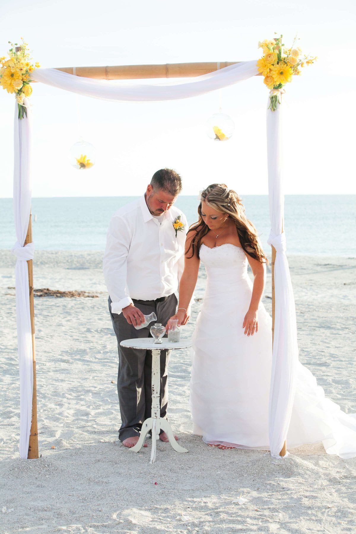 Wedding couple pouring sand together on a beach, under a bamboo arch with yellow flowers.