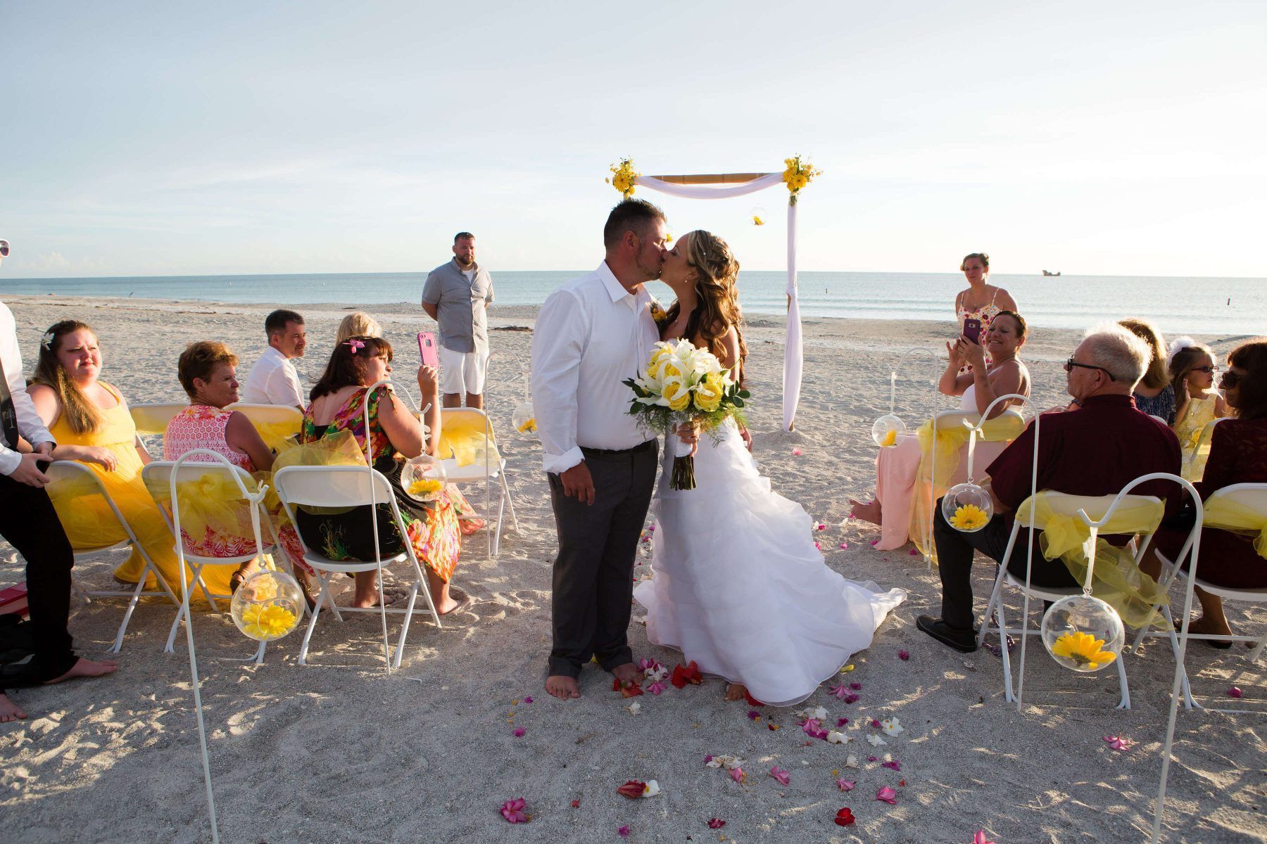 Couple kissing at a beach wedding ceremony, with guests seated on the sand, beneath a sunny sky.