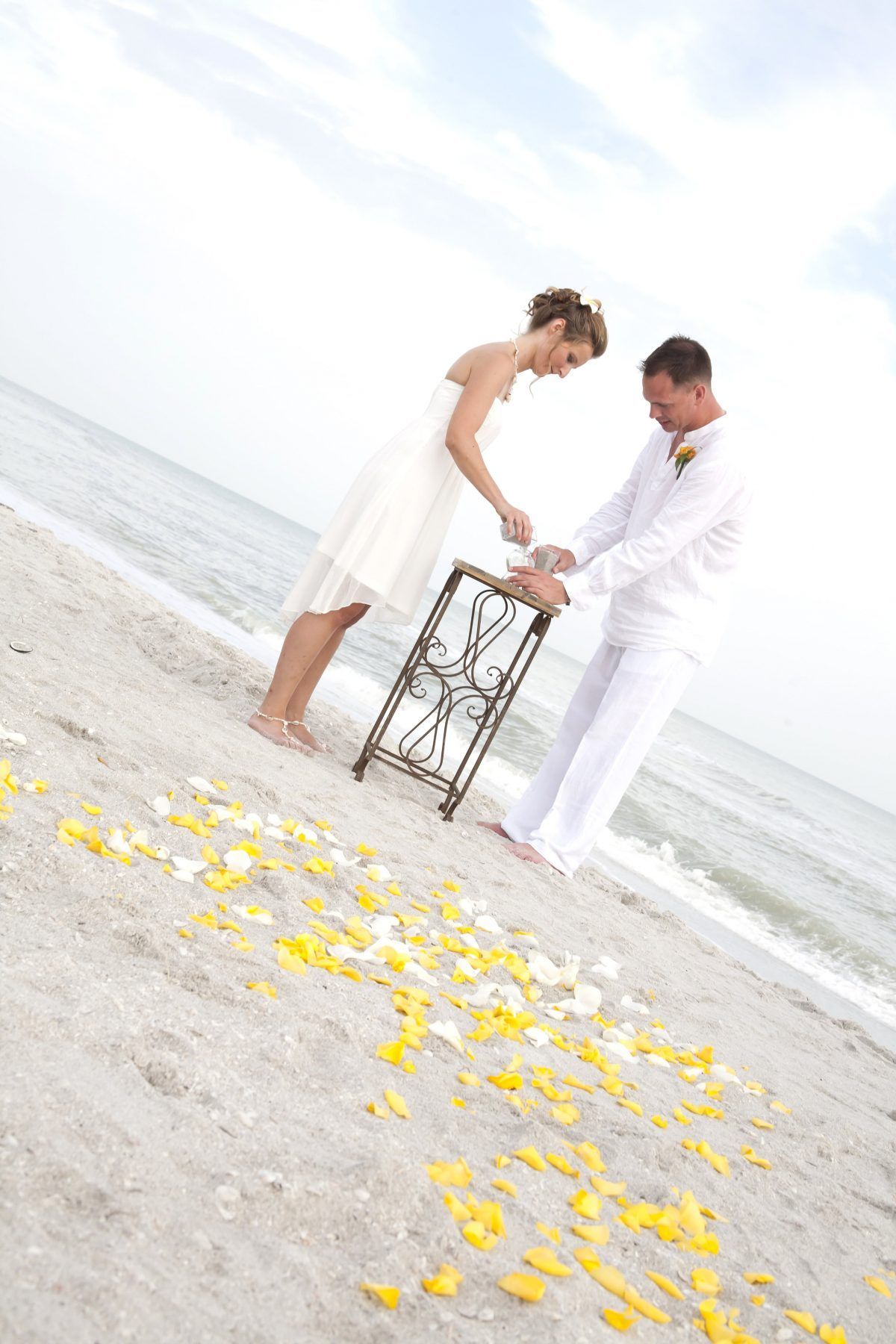 Bride and groom at beach wedding pouring sand into a unity vase, surrounded by flower petals.