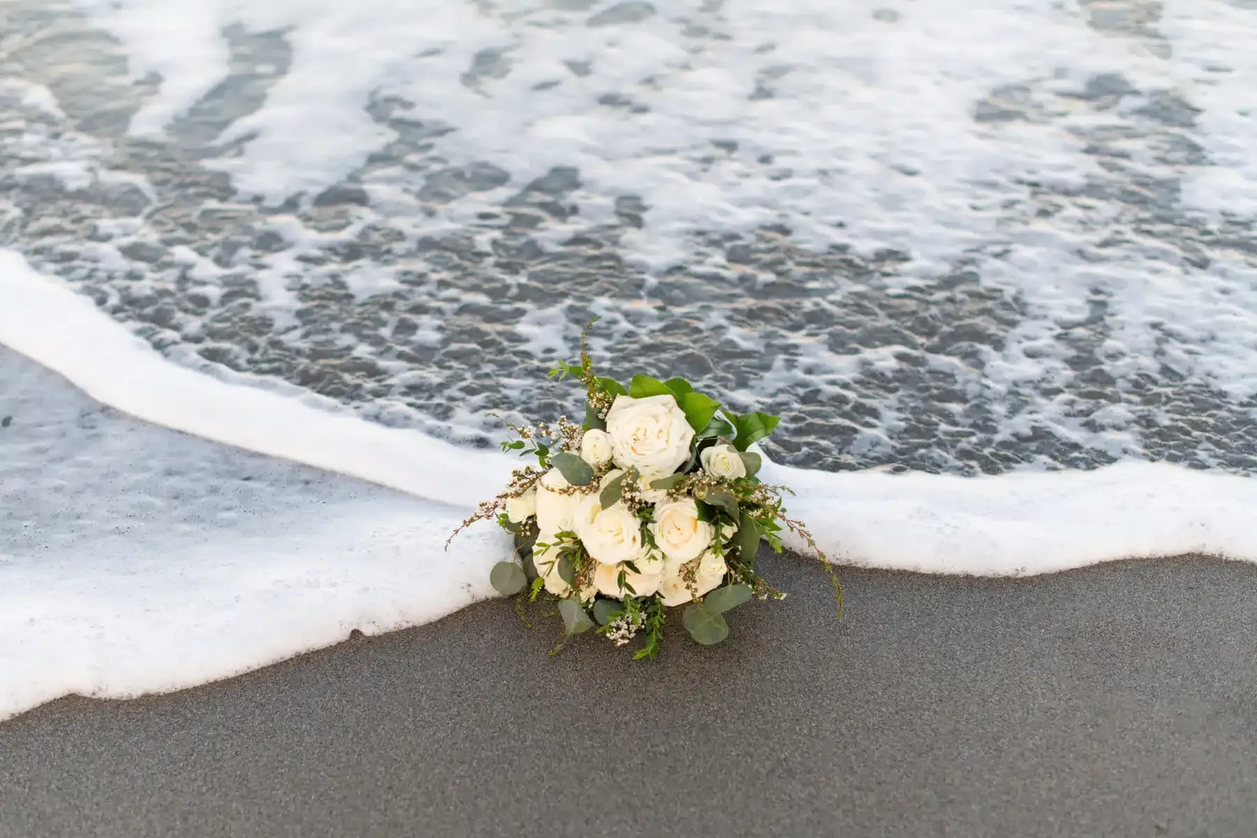 Bouquet of white roses and greenery on a sandy beach, wave cresting around it.