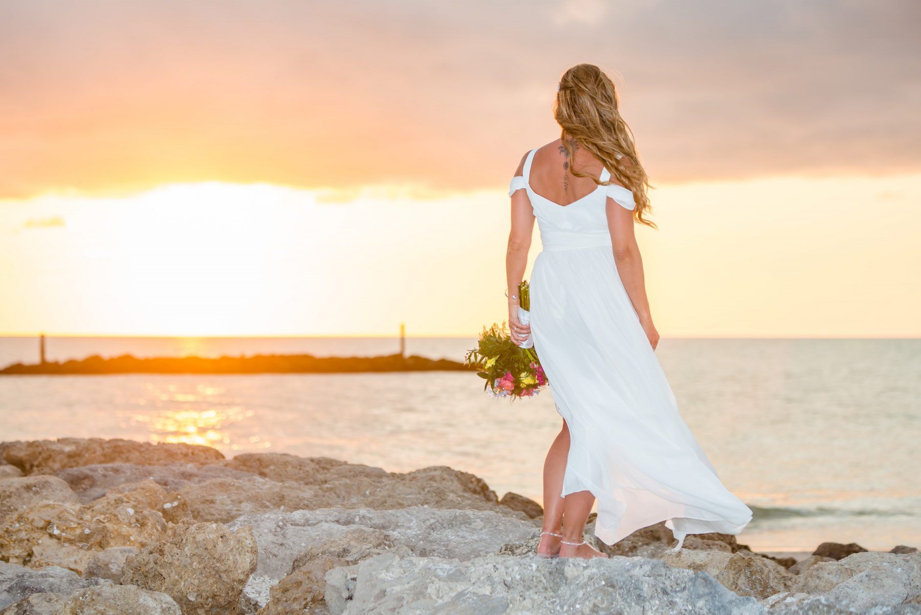 Woman in white dress with bouquet stands on rocks, facing sunset over the ocean.