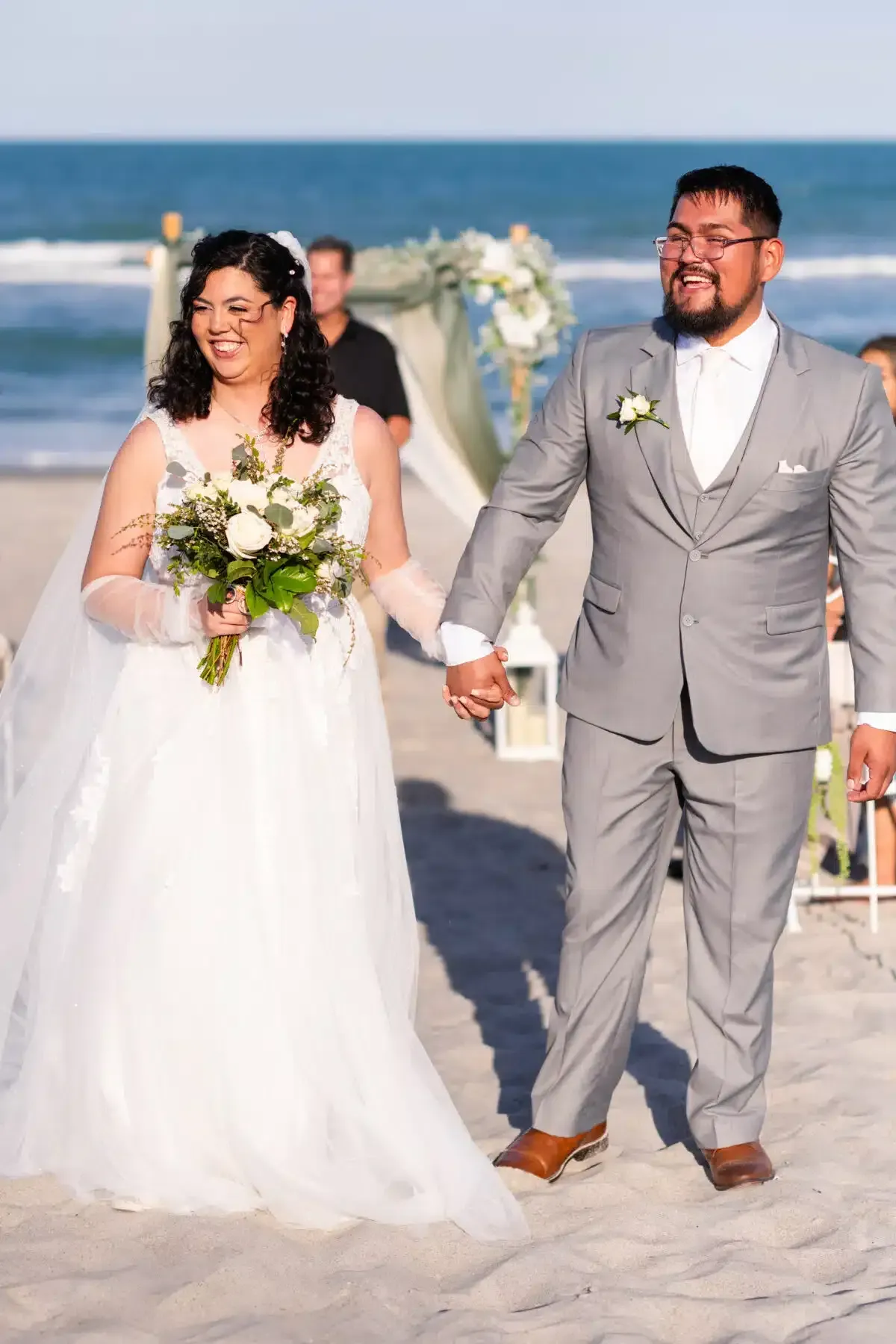 Newlyweds hold hands on a beach; woman in a white dress, man in gray suit, with ocean backdrop.