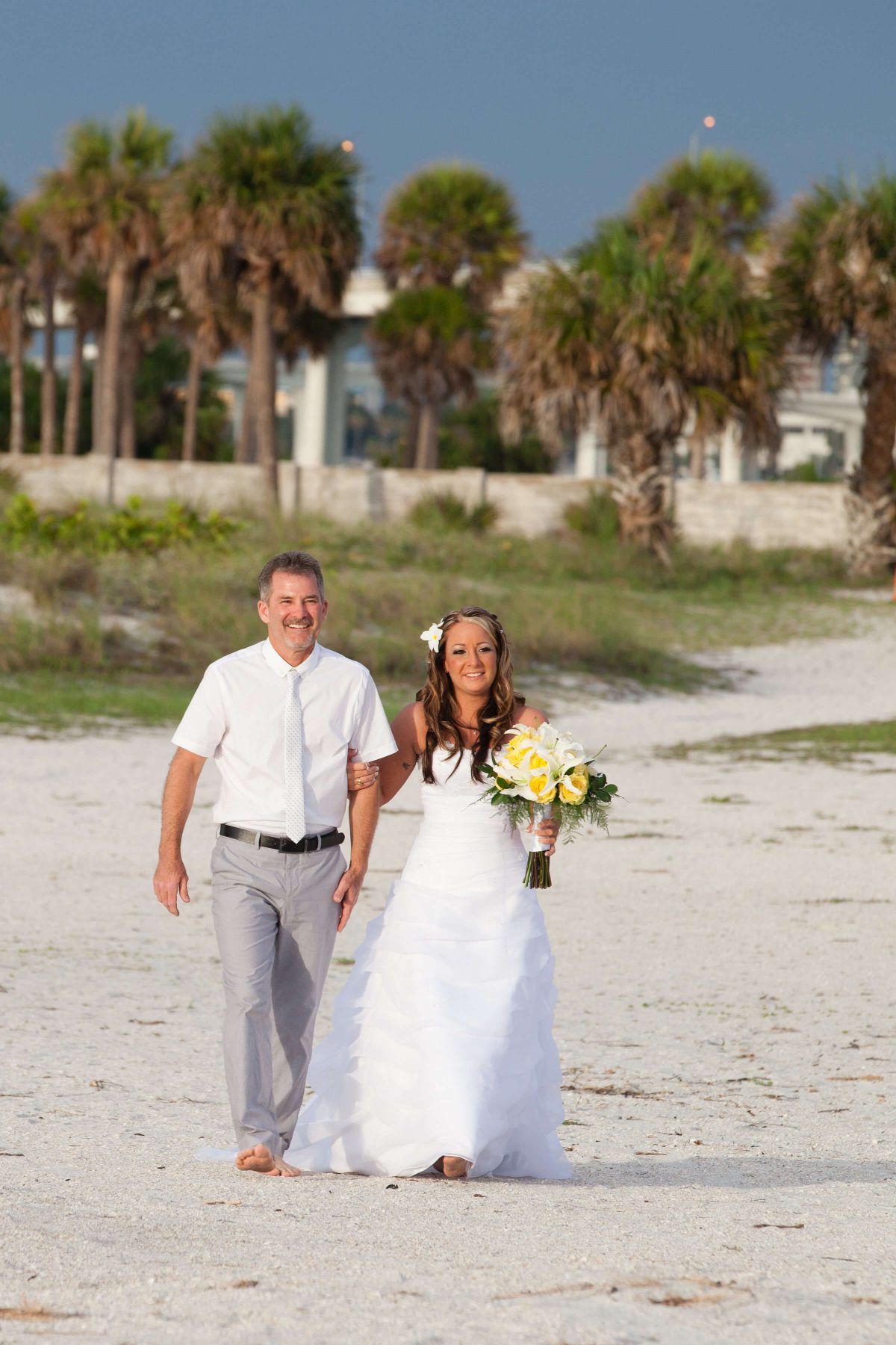 Bride and man walk on a beach. She wears a white dress and holds a bouquet, he wears light-colored pants and a button-up shirt.