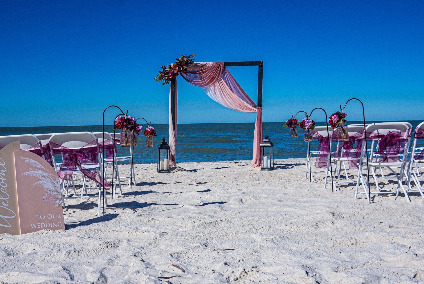 Beach wedding setup with arch, draped fabric, chairs, and ocean backdrop.