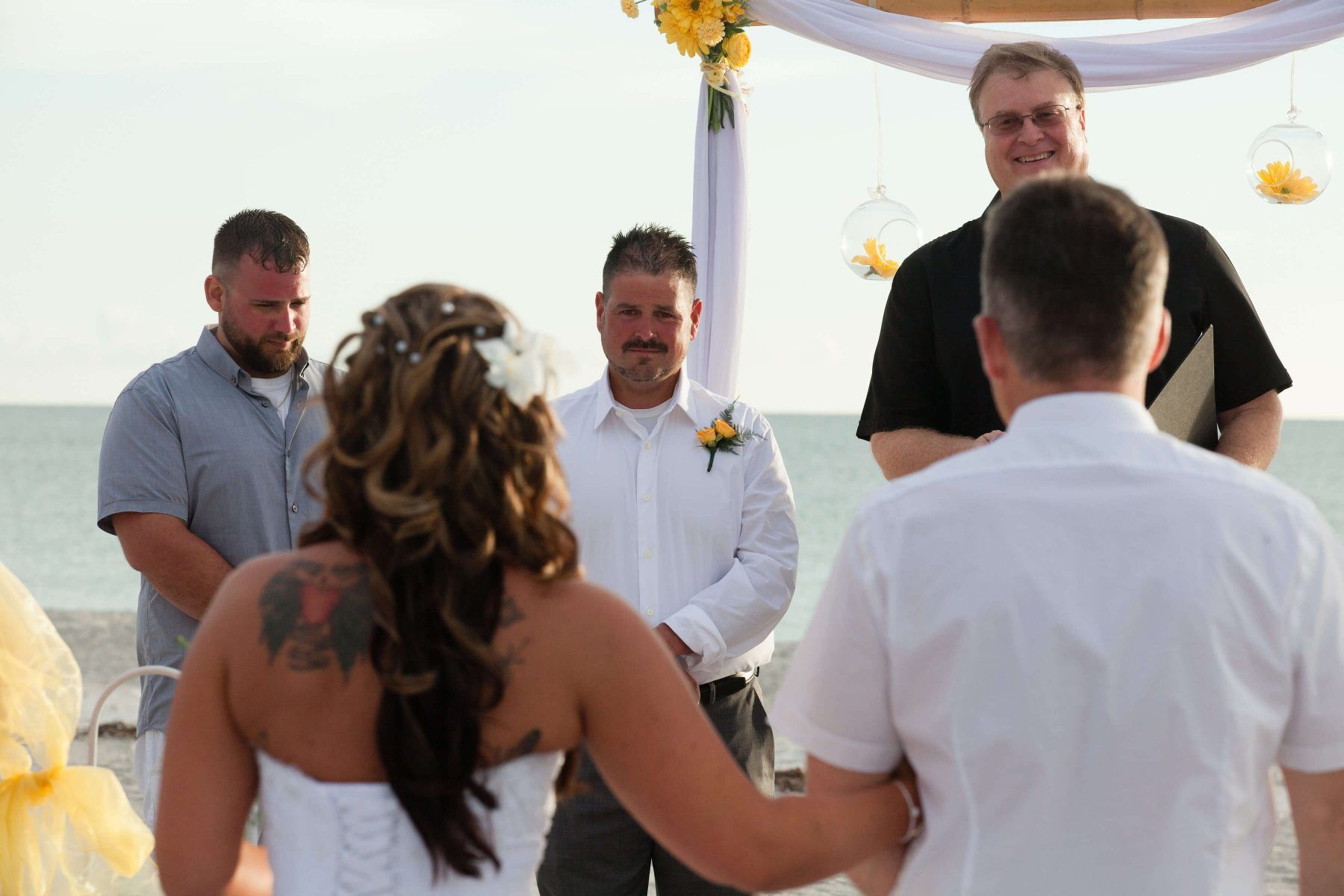 Wedding ceremony on a beach. Bride and groom face the officiant; guests stand nearby. Yellow and white decorations.