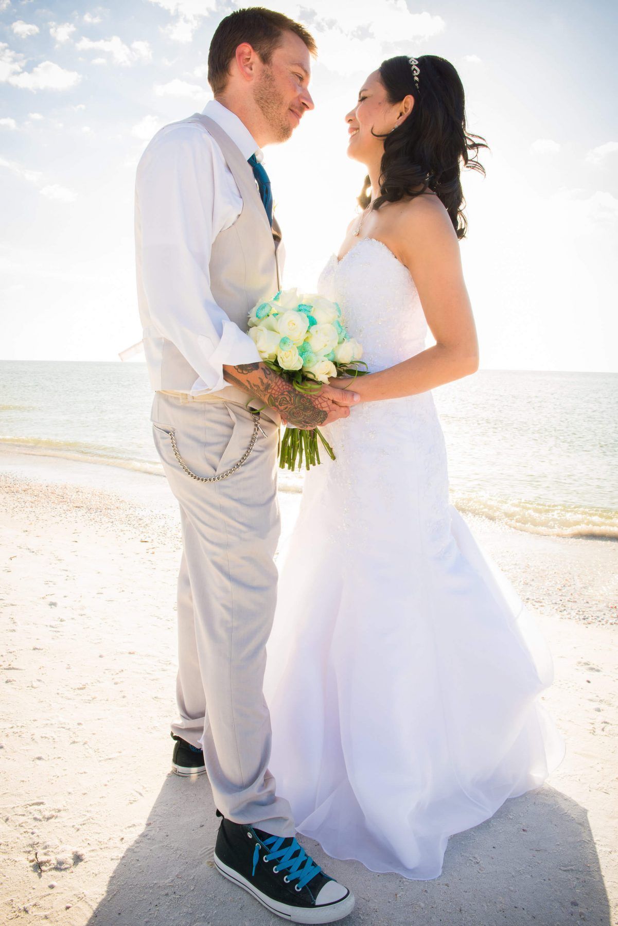 Couple on a beach on their wedding day, looking at each other. The bride in white dress, groom in suit with sneakers, holding flowers.